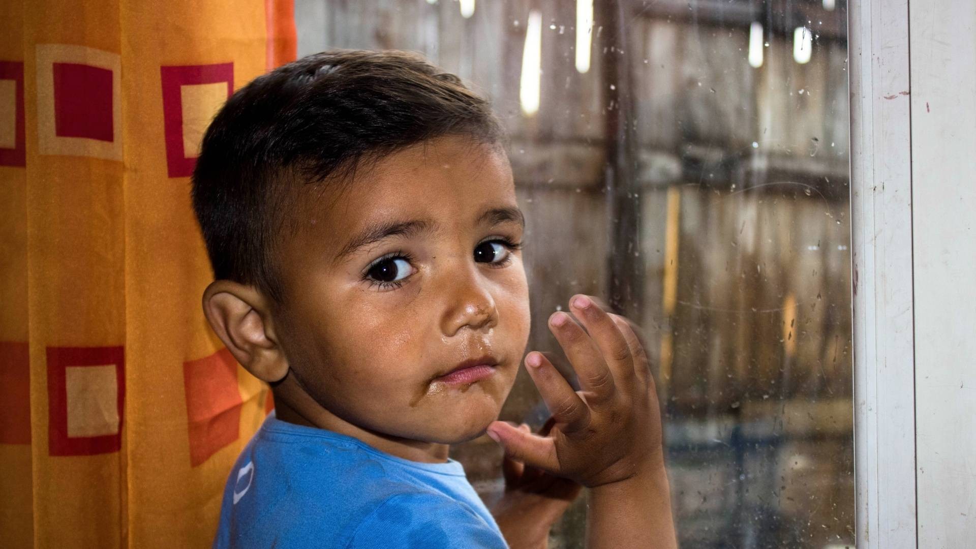 Serious boy in blue shirt looks back at camera, standing at a rain-covered window