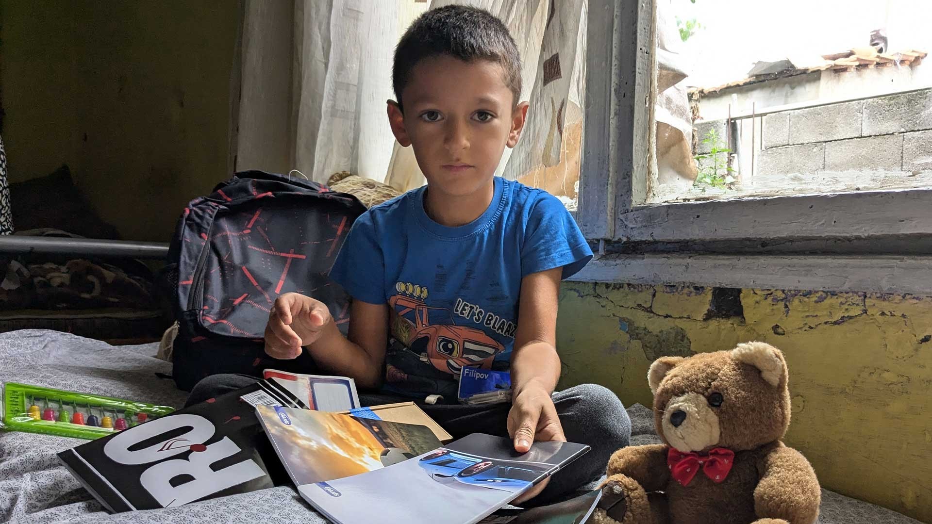 Young Roma boy in a blue T-shirt sits on a bed beside an open window. Behind him is a backpack, and in front of him, a teddy bear and school supplies.