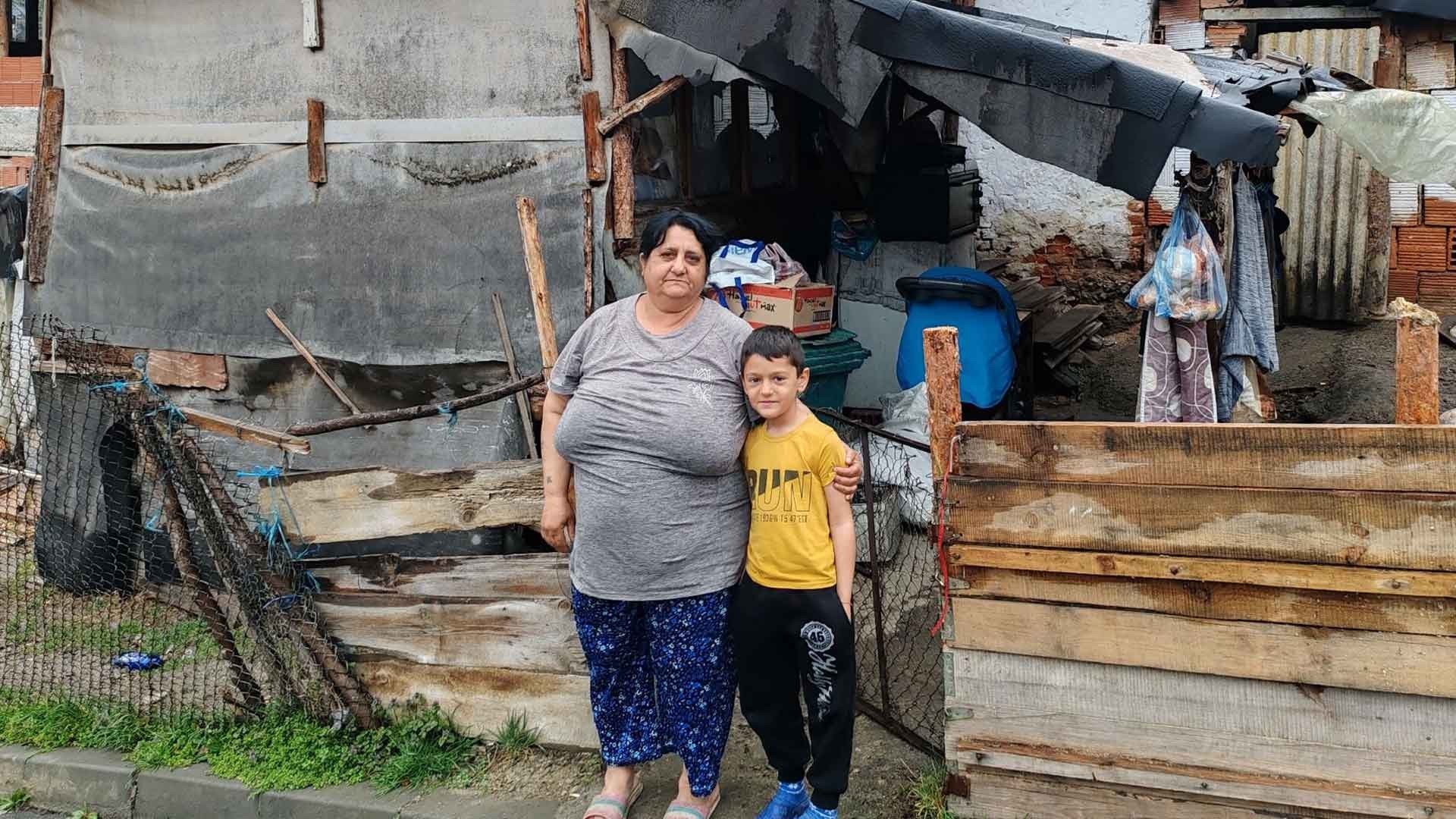 A young Roma boy in a yellow T-shirt stands embraced by an older Roma woman in front of a dilapidated house.