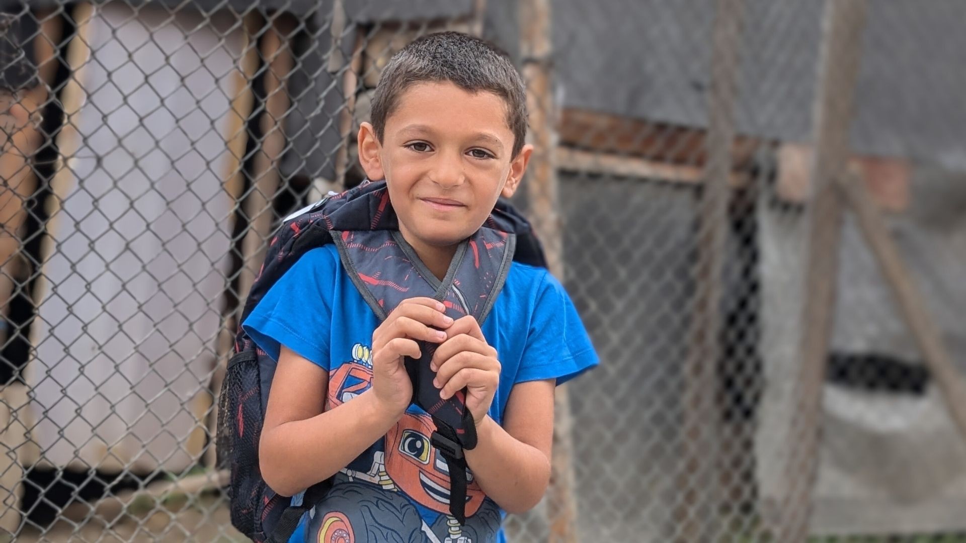 Young Roma boy standing outside a wire fence, smiling shyly as he wears his new backpack.