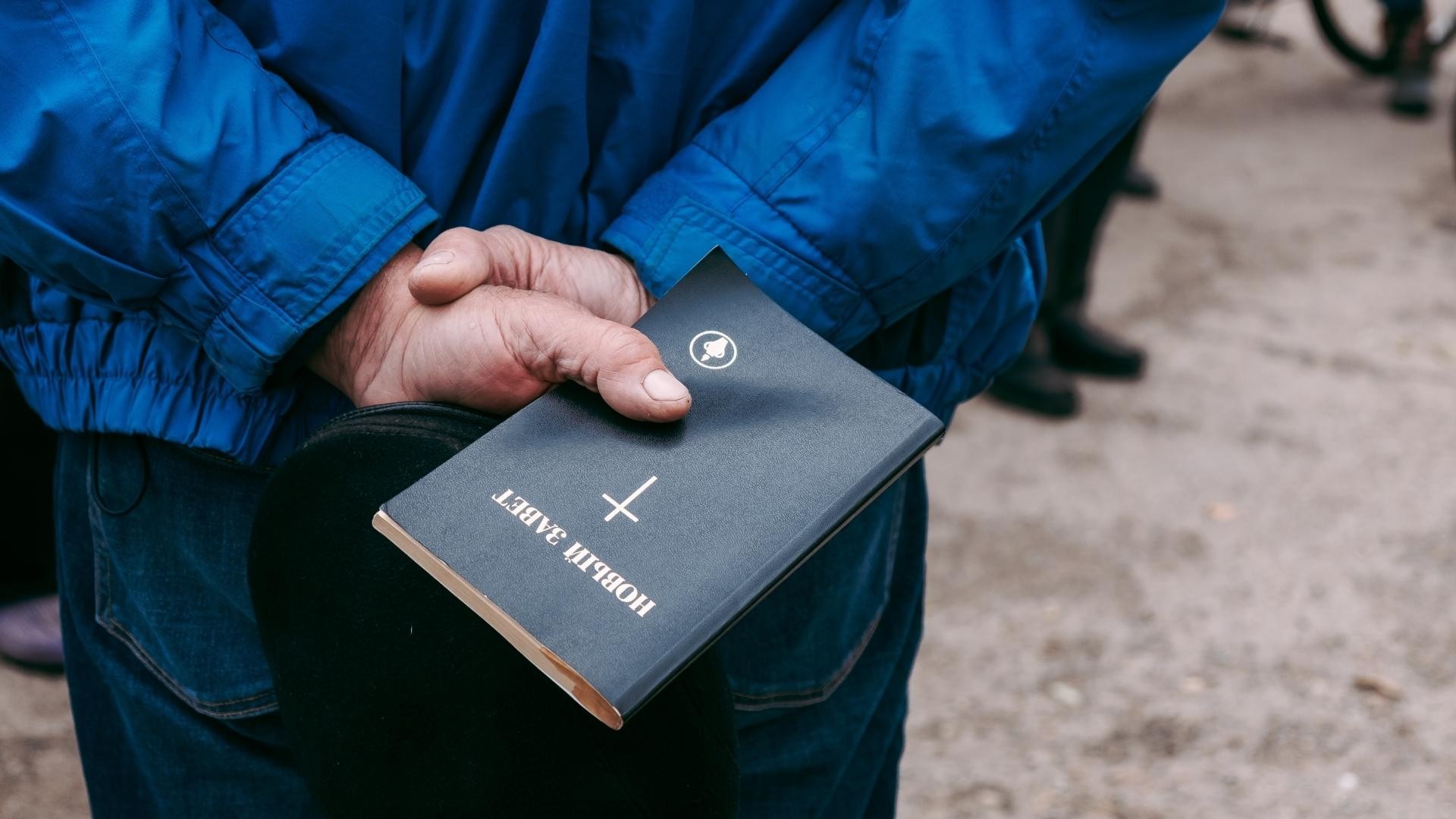 Close-up of a man's hands holding a hat and Bible behind his back in a peaceful stance.