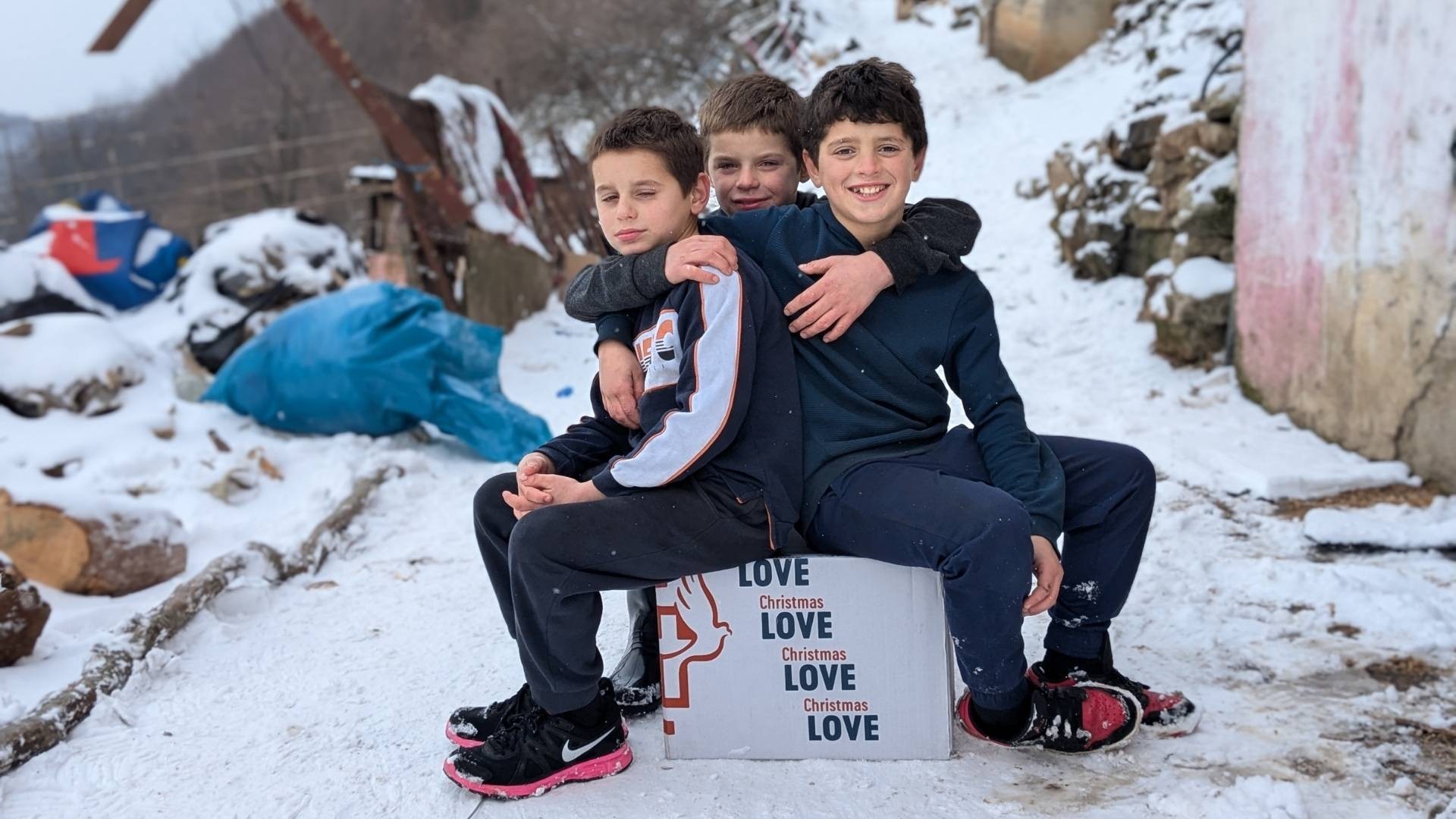 Three young boys sitting on a box in the snow, holding onto each other and smiling.