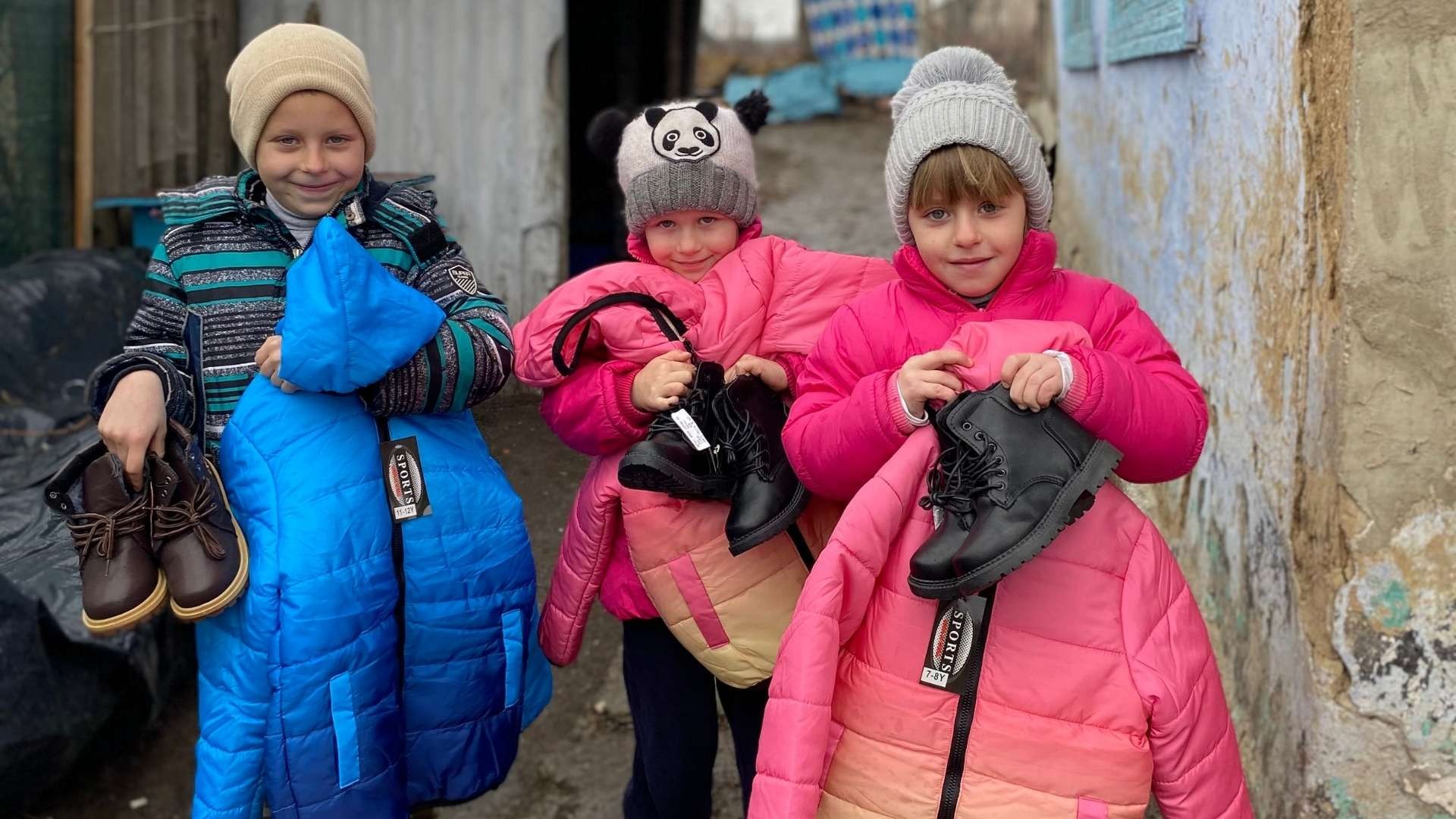 Three smiling children proudly holding their new winter jackets and shoes.
