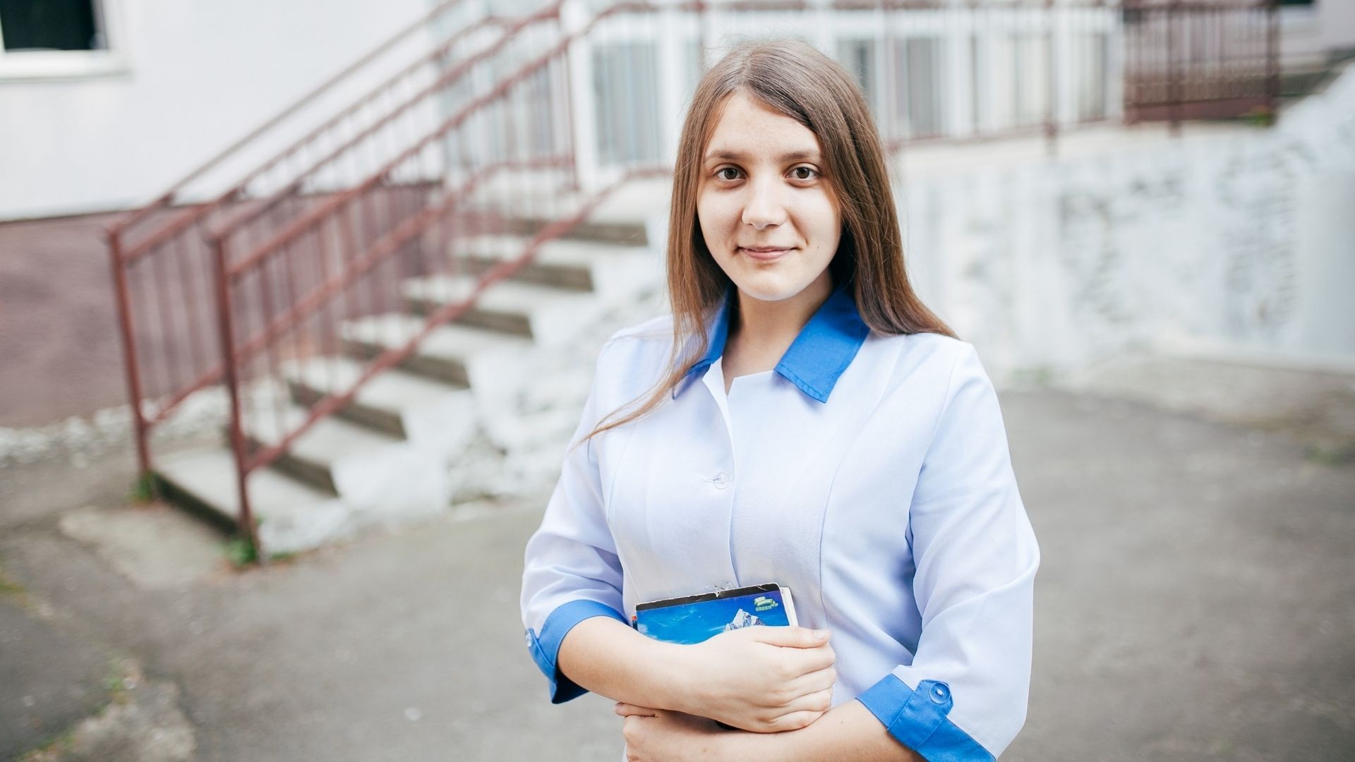 Young woman in a nurse’s uniform, smiling shyly while holding a notebook close to her chest.