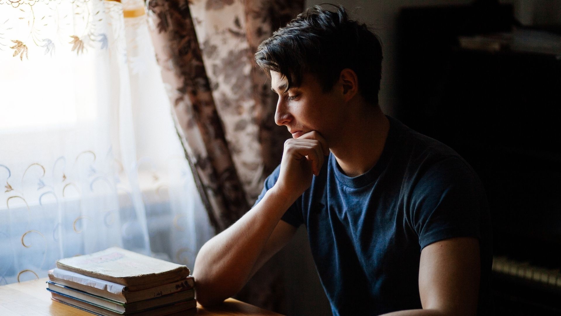 Young man sitting at a desk by a window, looking thoughtfully down at his books.