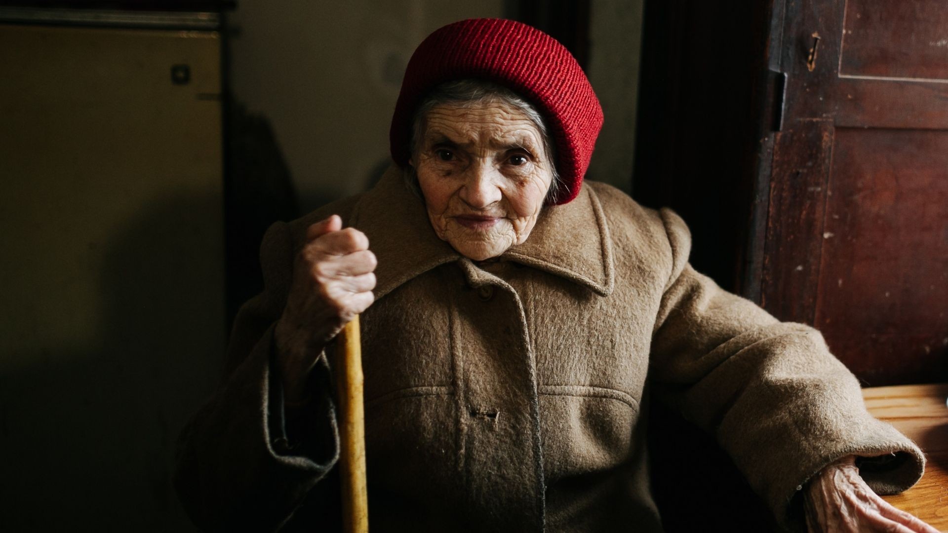 Elderly woman in a red knitted hat and winter coat sits at a table indoors, holding her cane.
