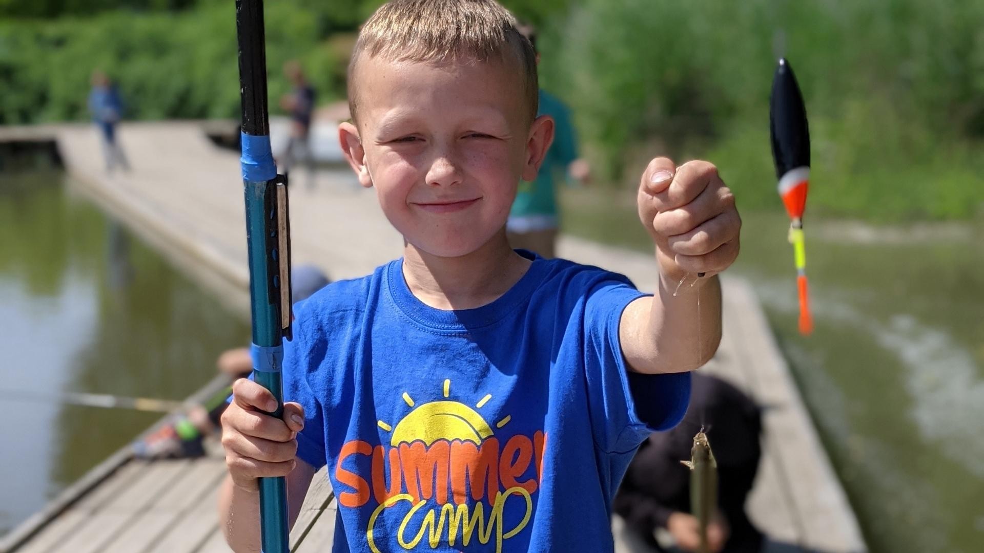 Fair-haired boy in a blue summer camp t-shirt proudly holding up his fishing rod with a fresh catch.