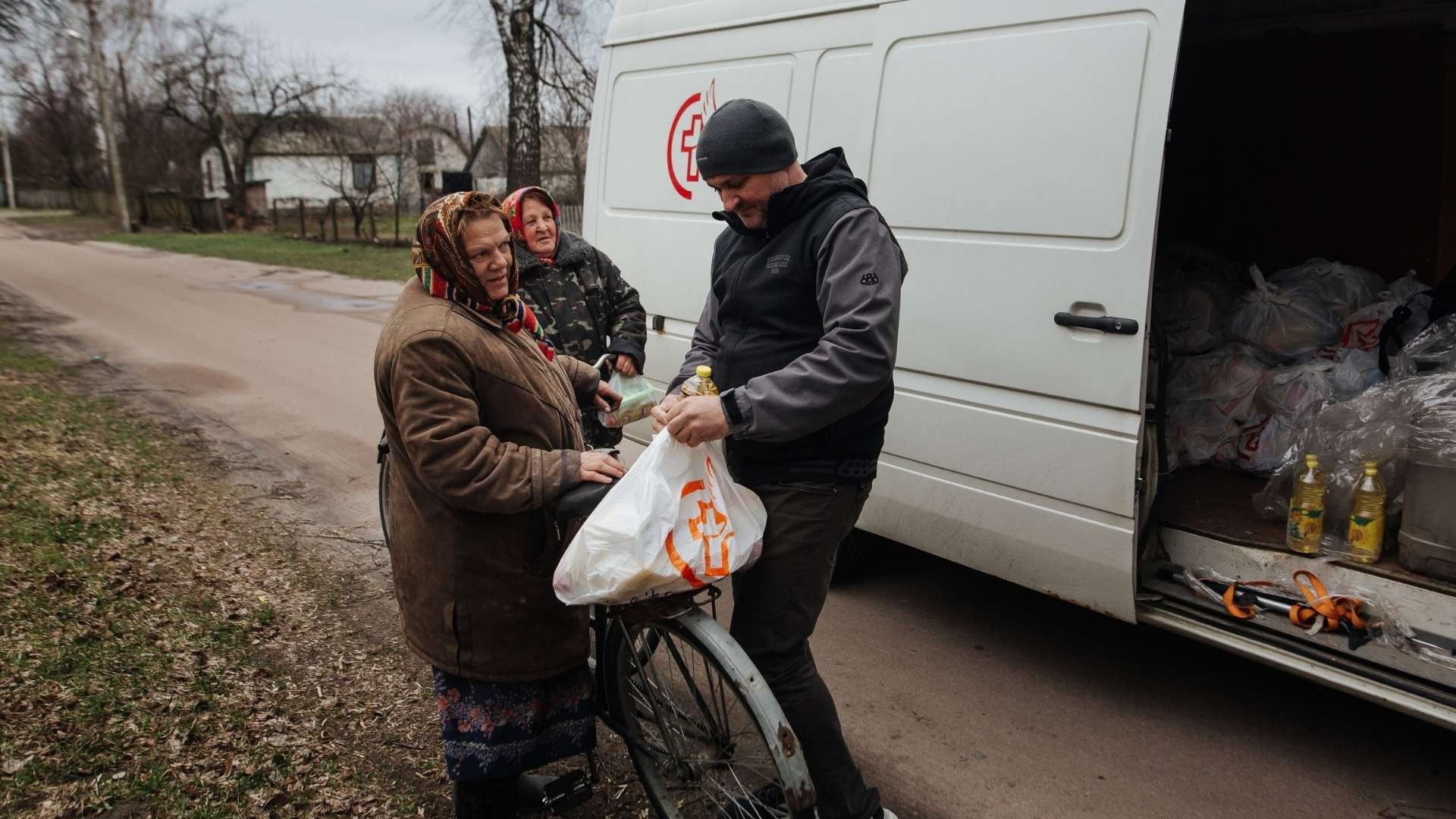 Young man tying a grocery bag to a bicycle, sharing a moment with two elderly women bundled in layers of winter clothing.