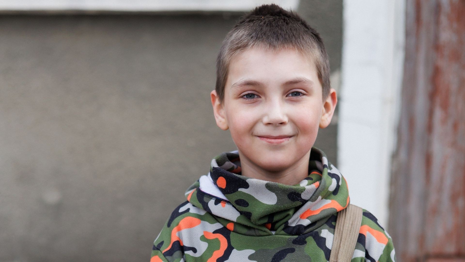 A young boy in a camo sweater stands outside, smiling.