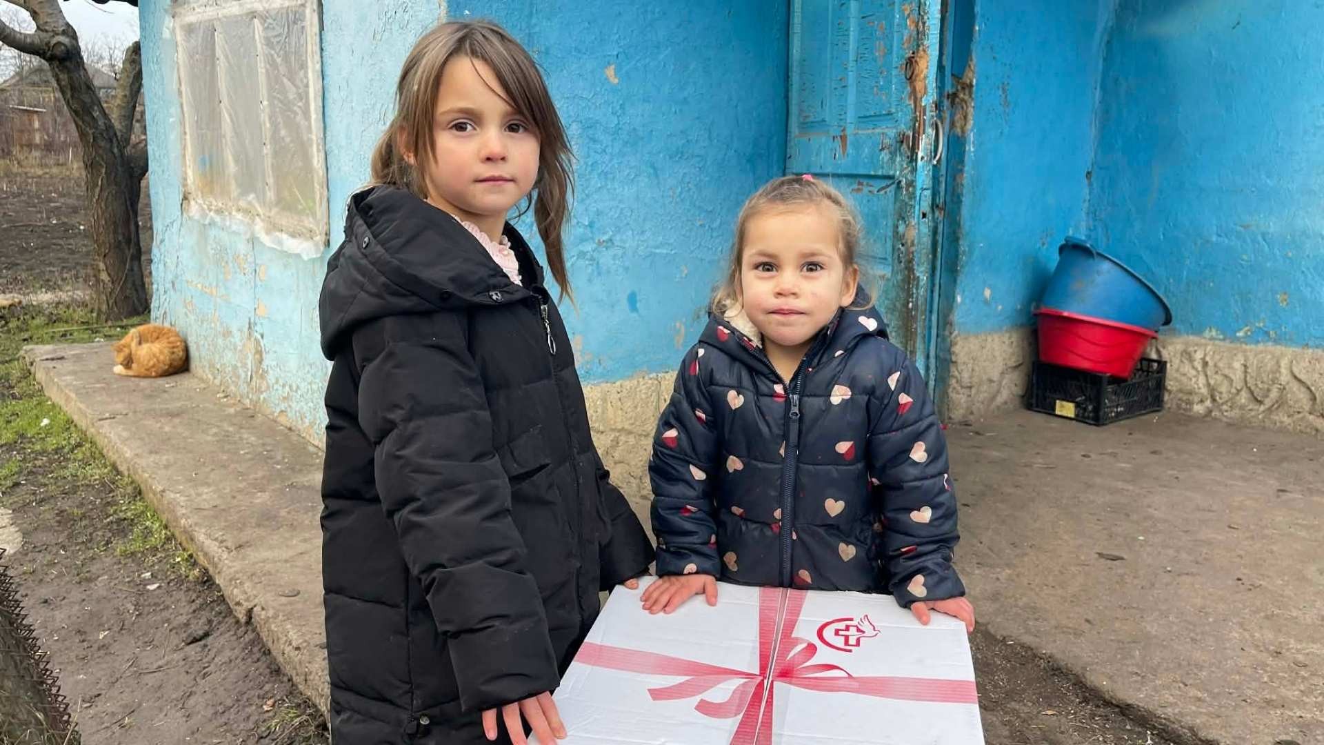 Two girls stand beside a large Christmas box outside their home.