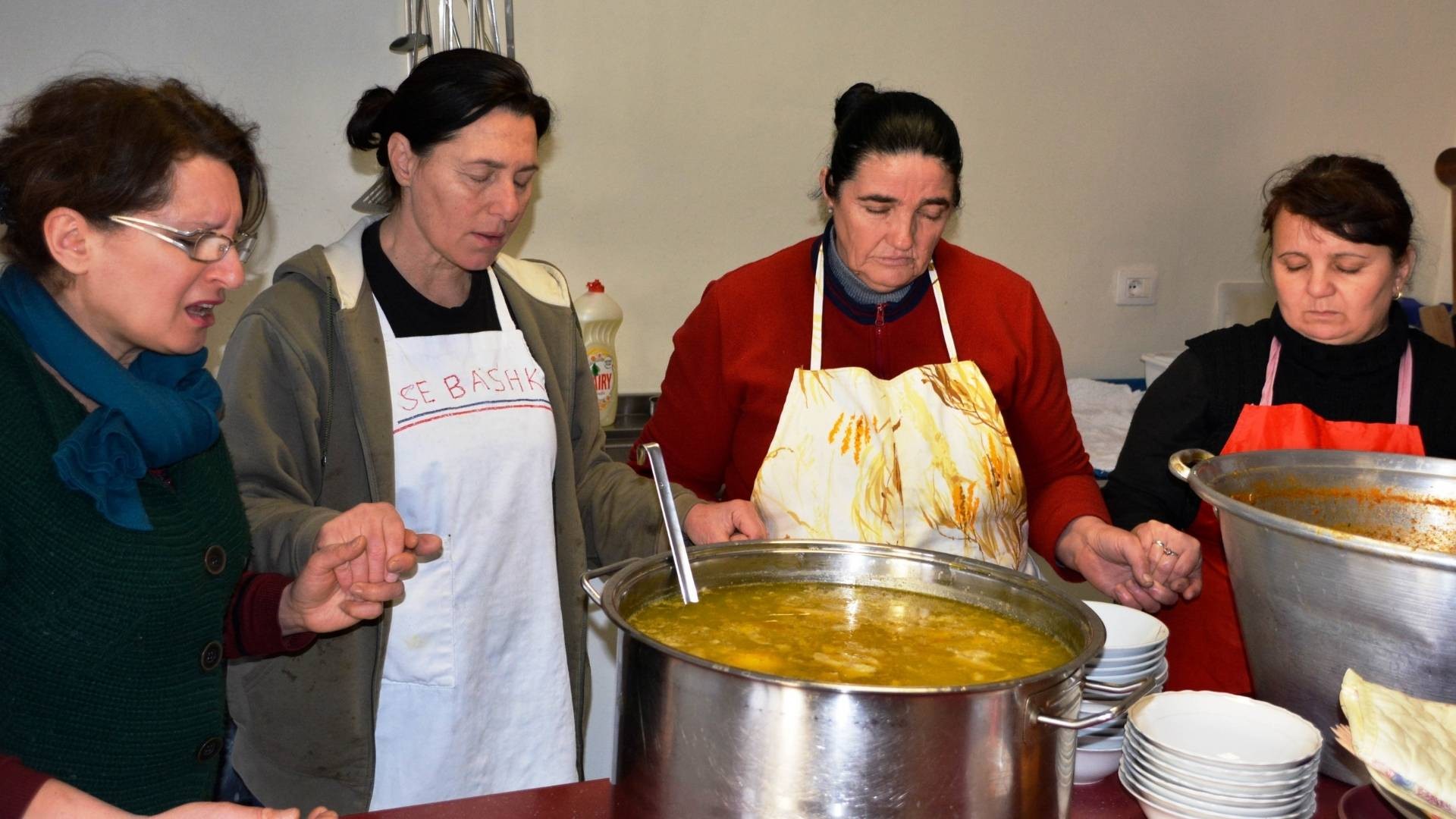 Four women in aprons pray together before serving meals at a soup kitchen.
