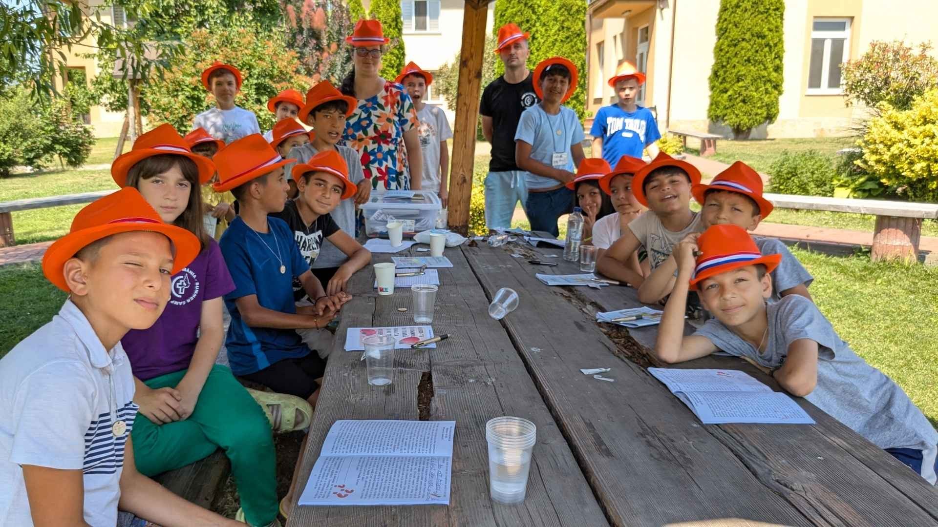 Teens in bright orange hats relax together around a wooden deck table at camp.