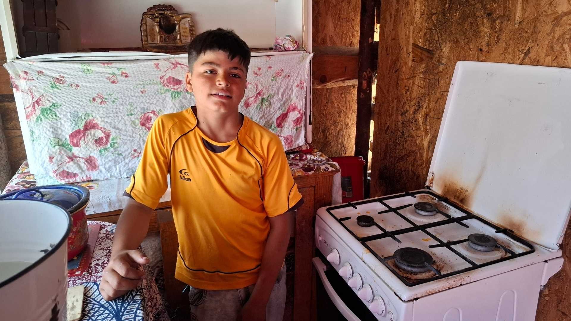 Teen boy in orange shirt stands in cramped kitchen with gas stove and water cans.