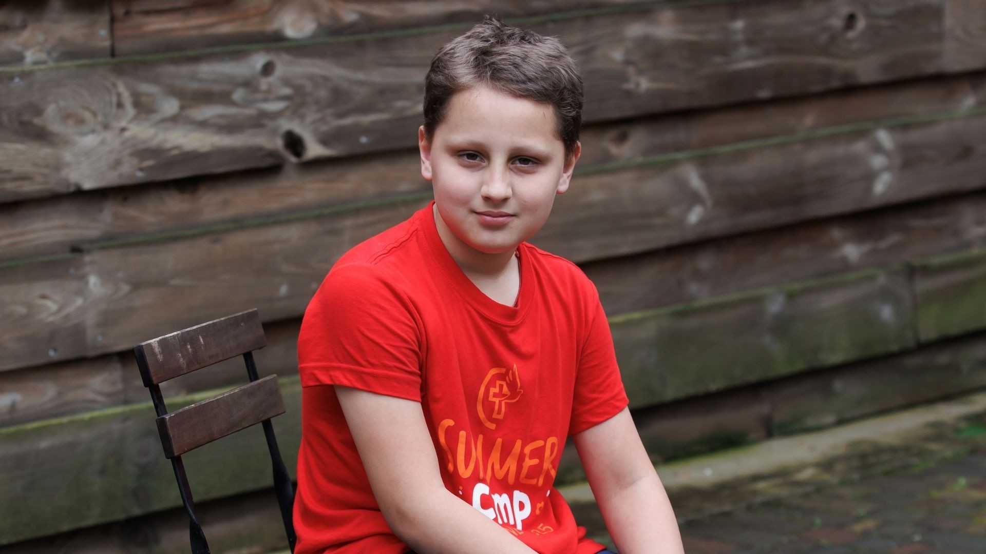 Boy in red camp shirt sits alone by plank wall with a faint smile
