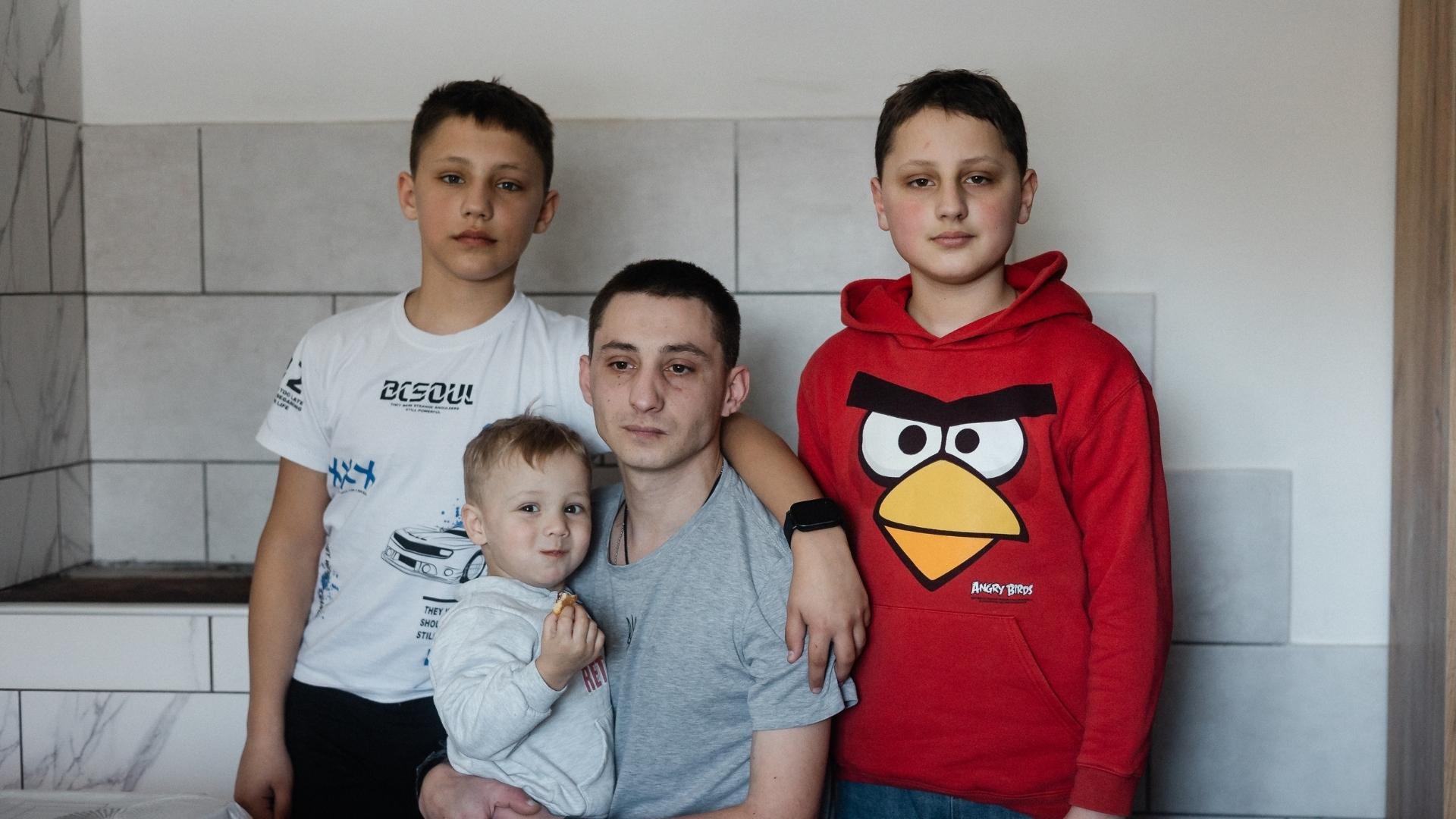 Somber father holds child as two boys stand close, one hugging his shoulder.
