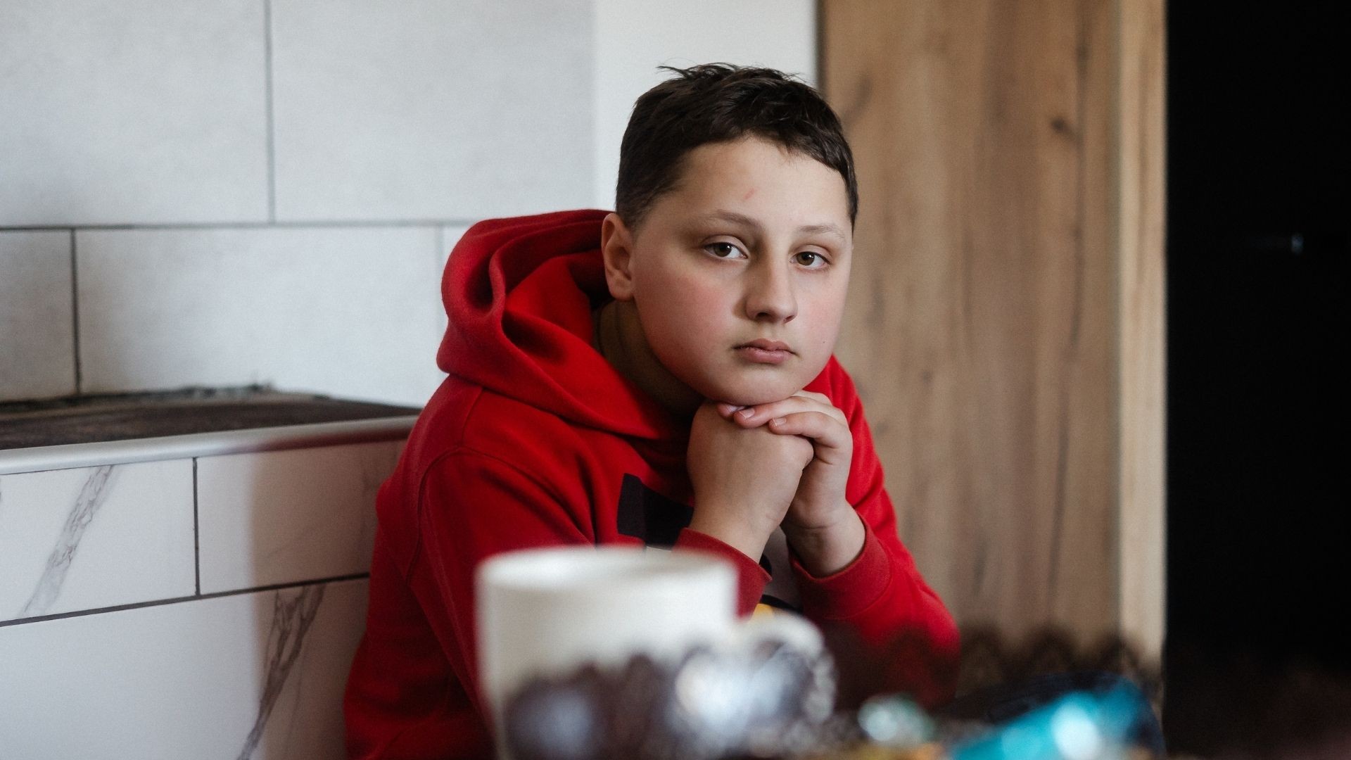 Sad young boy rests his head on folded hands while sitting in the kitchen