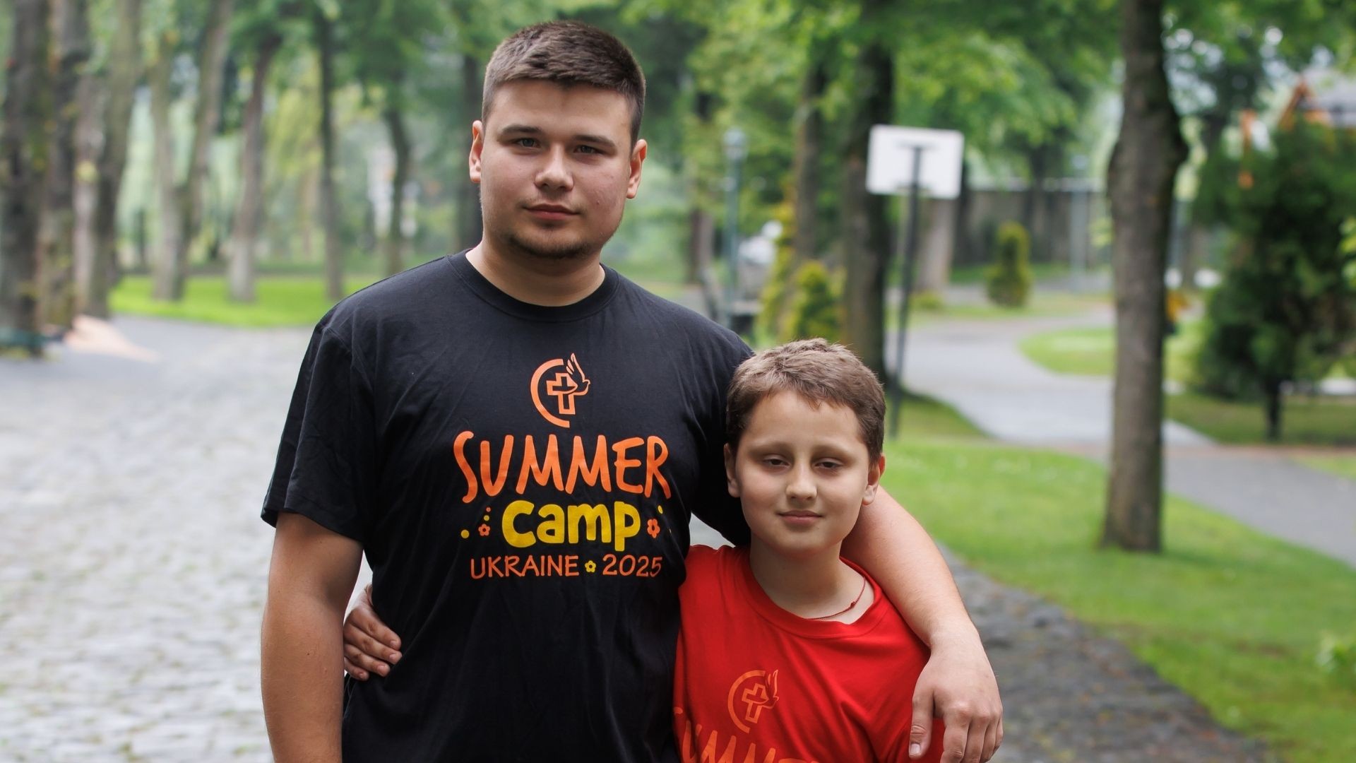 Boy and young camp leader stand arm in arm outdoors at summer camp.