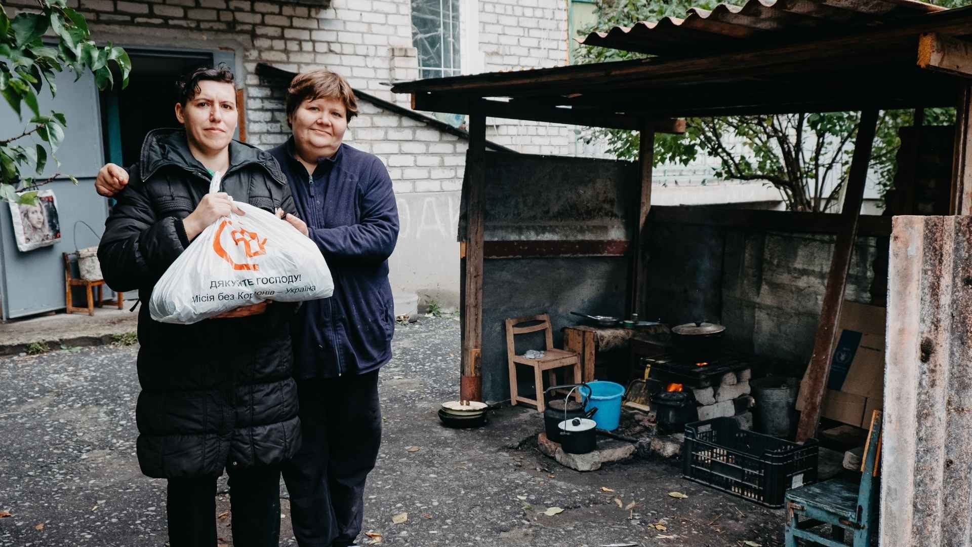 Two women stand outside with a bag of groceries as a pot cooks on a makeshift stove behind them.