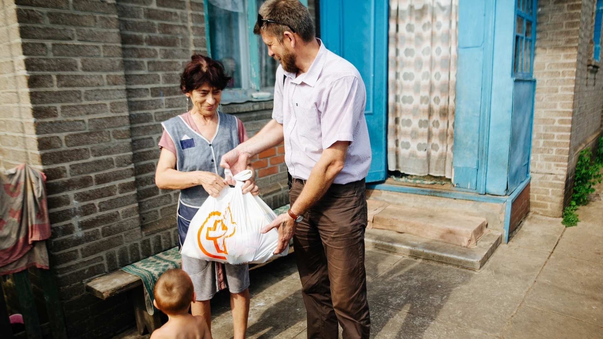 Bearded man handing a full grocery bag to a woman in an apron, with a small child nearby.