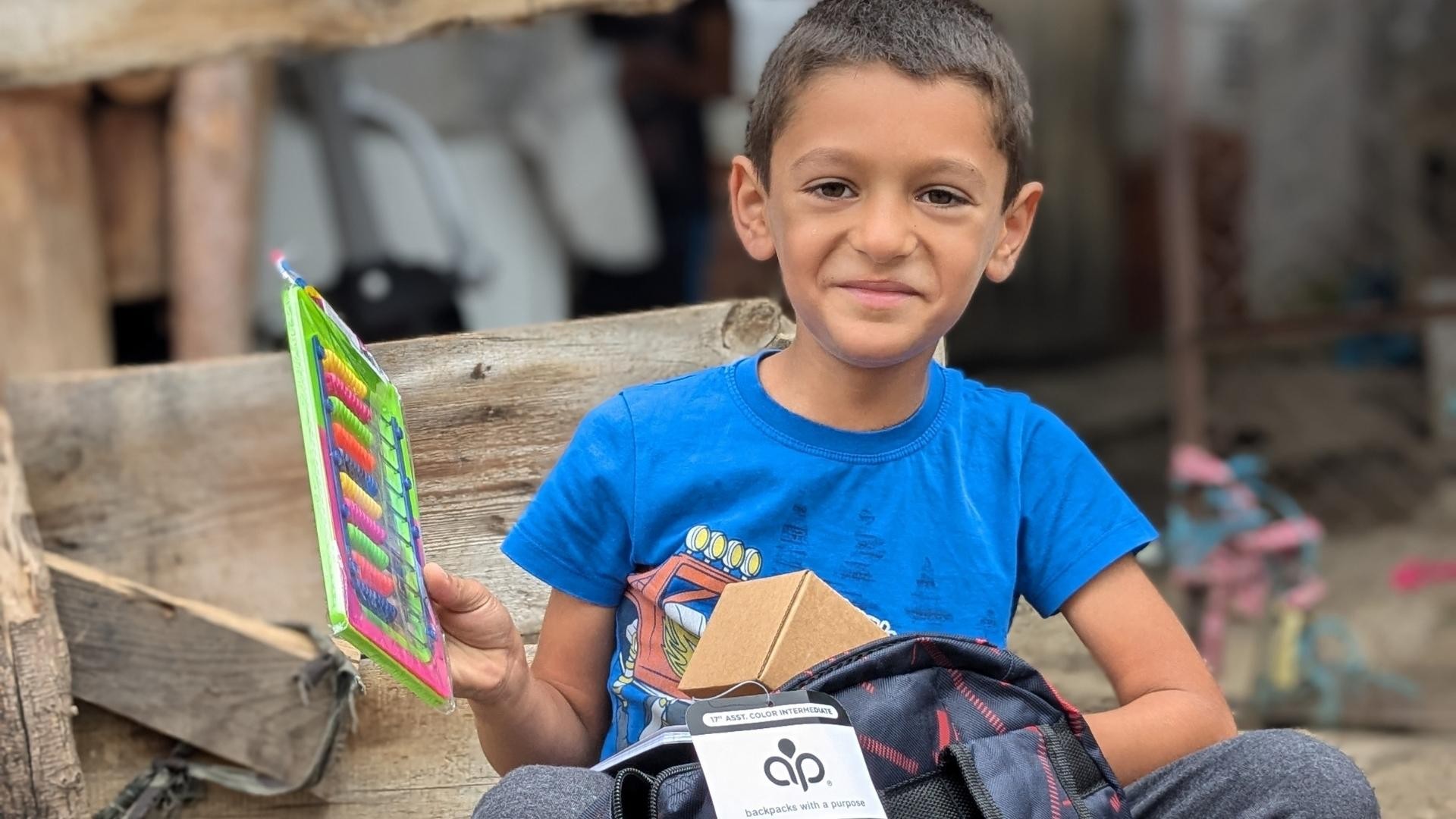Young Roma boy smiling while sitting on an outdoor step, holding his new school supplies.