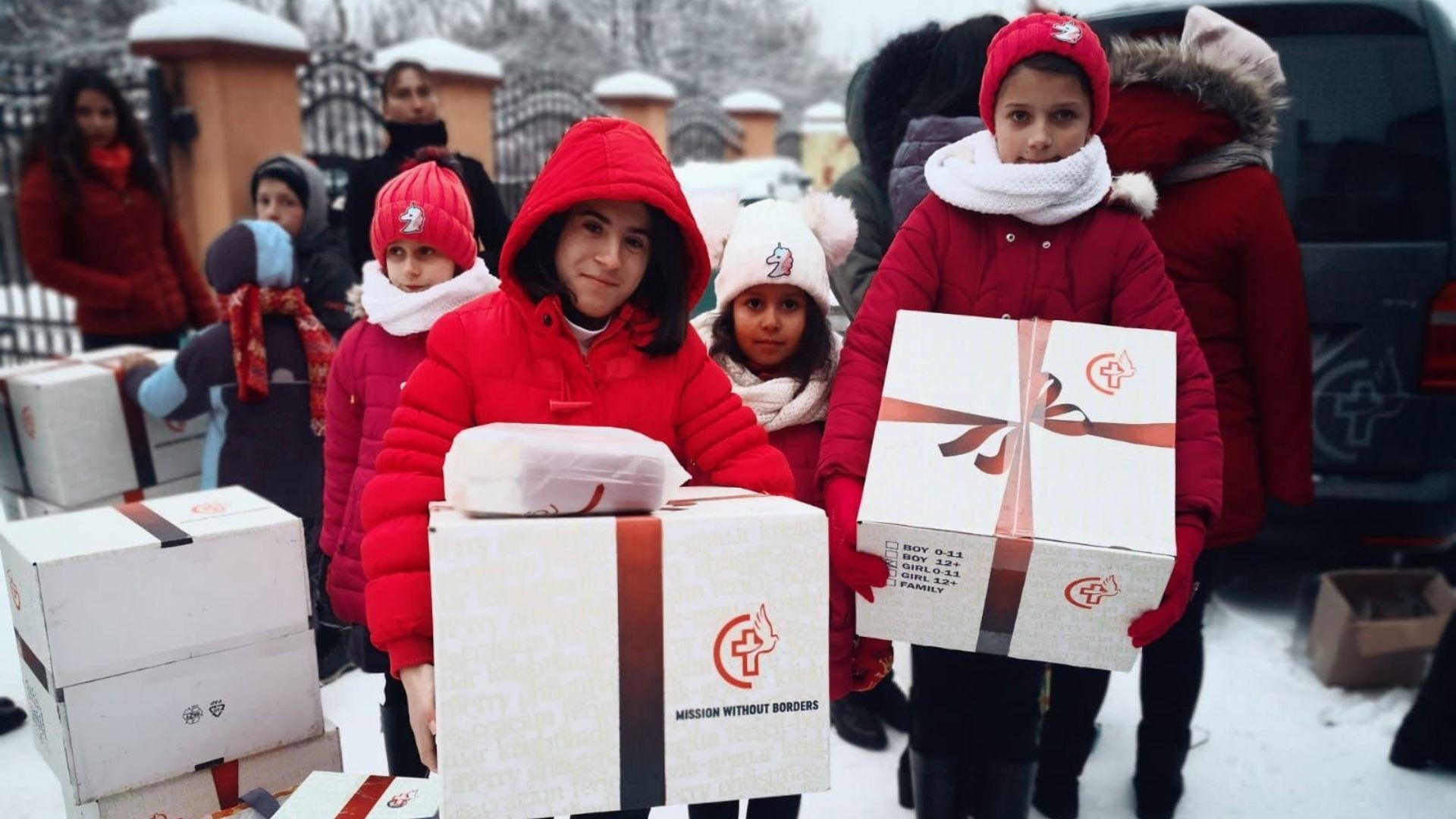 Children in red winter jackets and knitted hats stand in the snow holding large Christmas gift boxes, with a delivery vehicle and more stacked boxes visible in the background.
