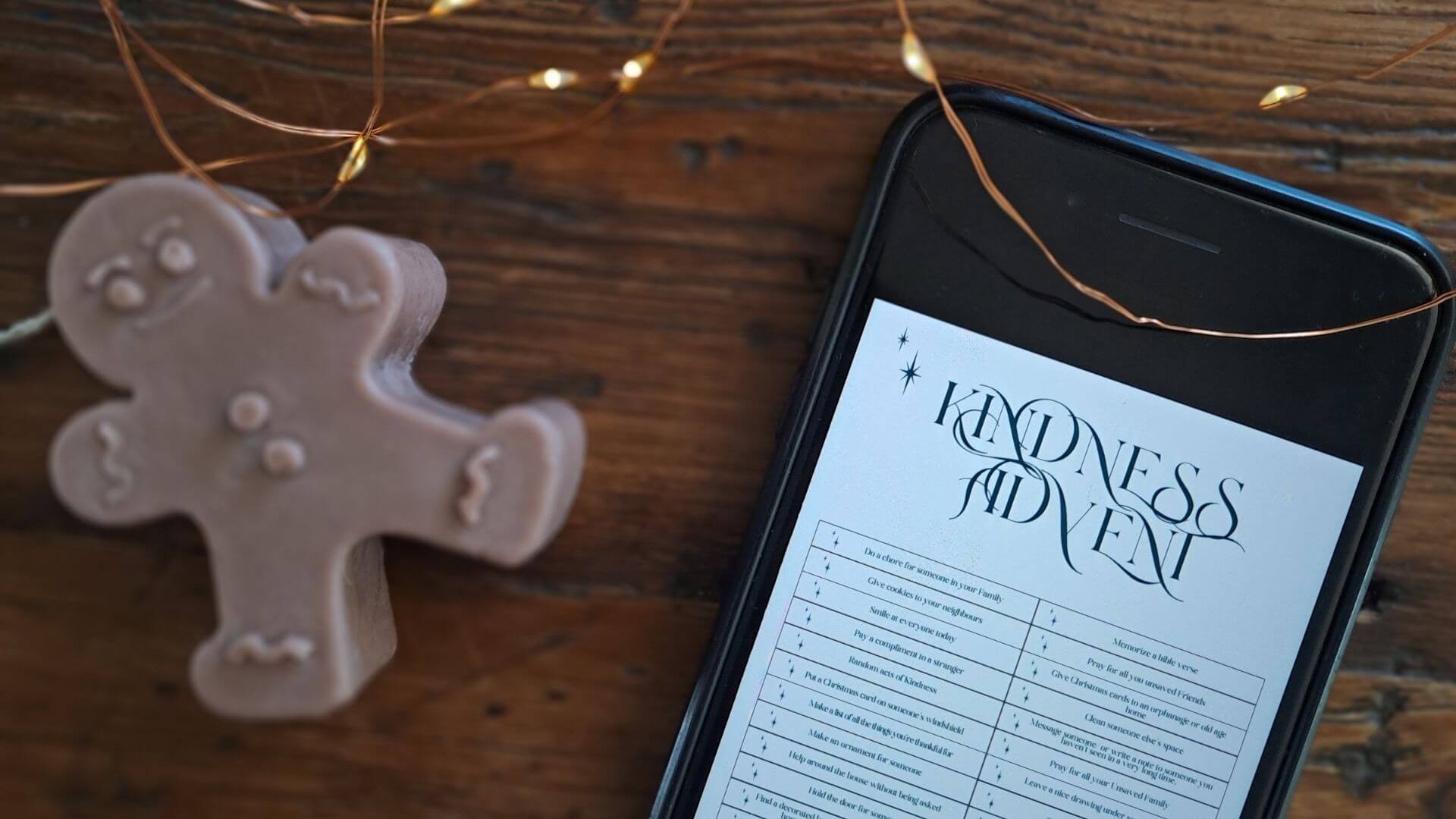 Mobile phone showing Advent list on wooden desk with Christmas lights and gingerbread candle.