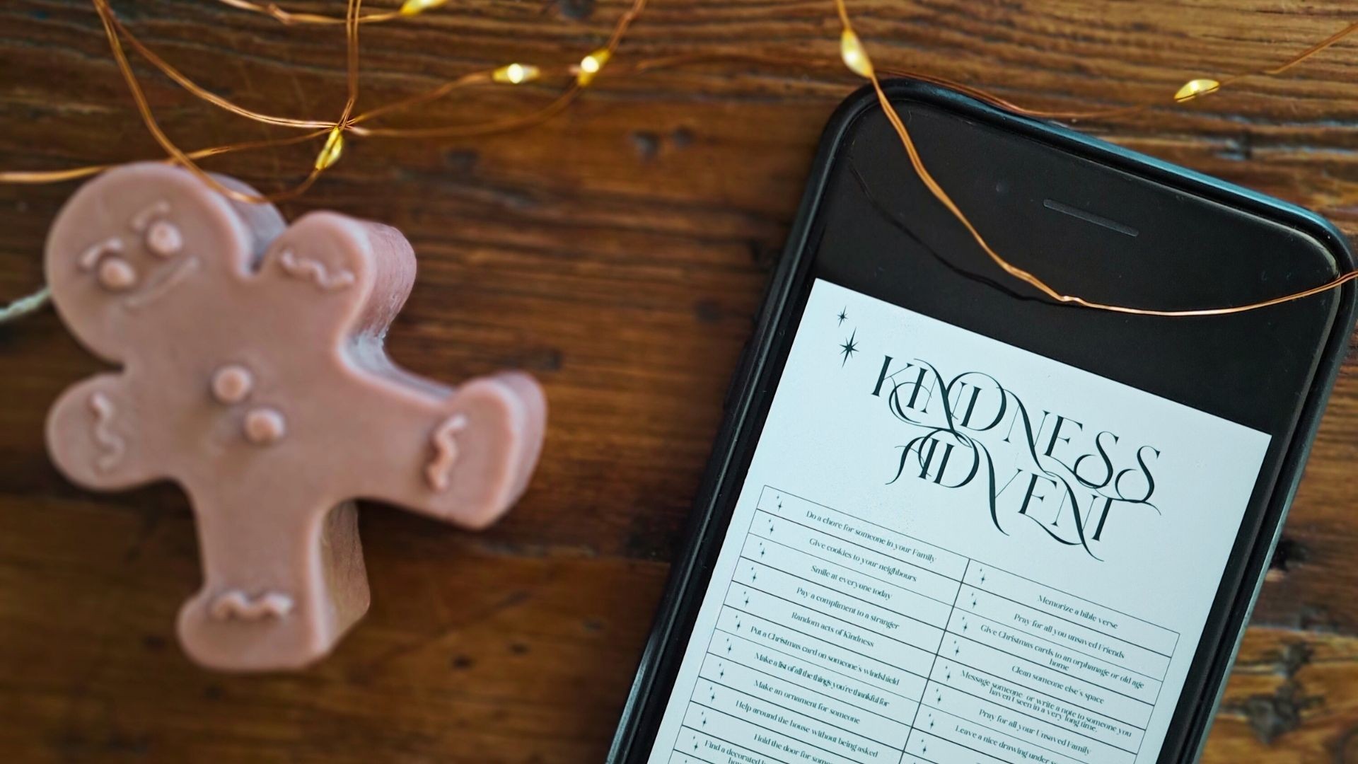 Mobile phone showing Advent list on wooden desk with Christmas lights and gingerbread candle.