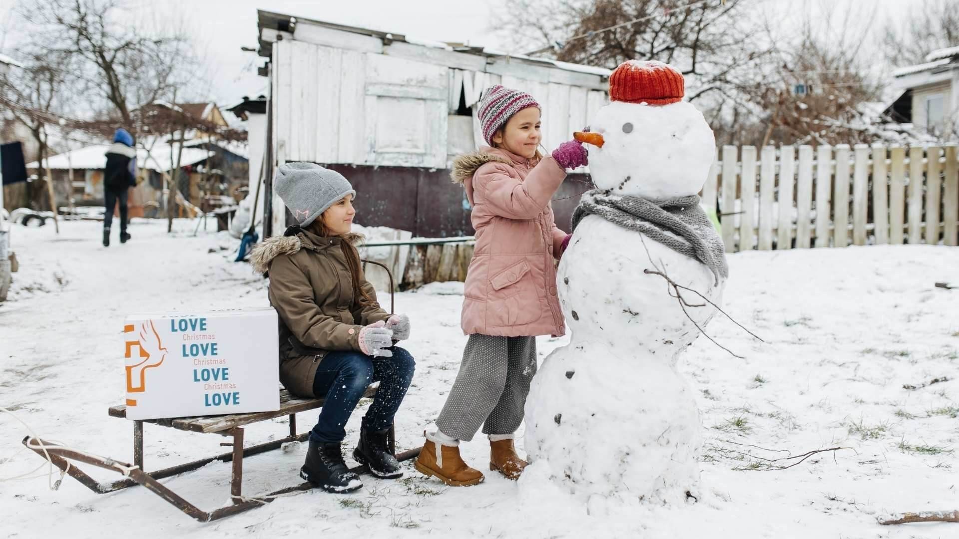 Two young children play in the snow — one adds an eye to a red-capped snowman while the other sits on a sled next to a Christmas gift box.
