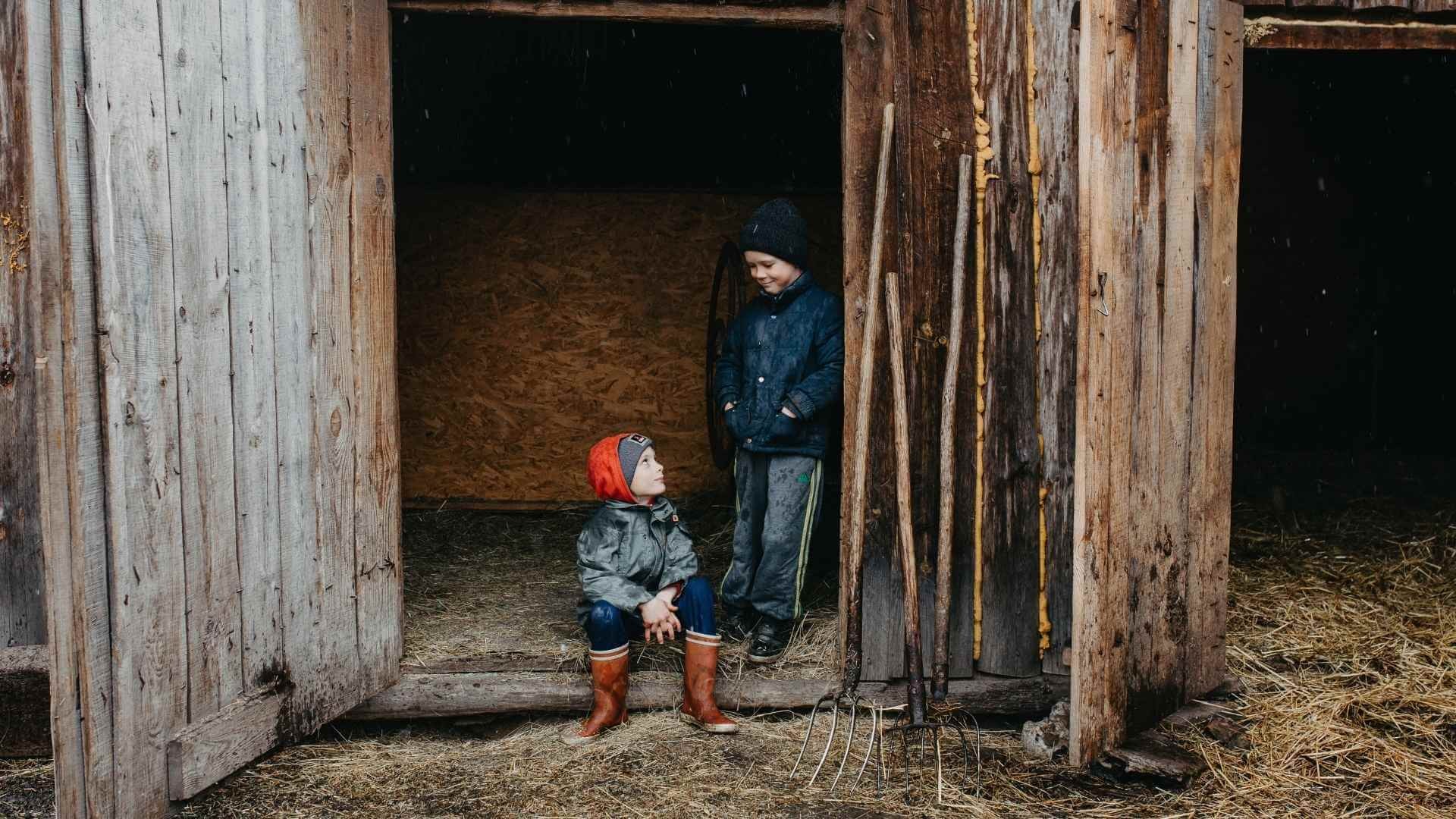 Two boys stand in a barn doorway, sharing a quiet moment together with pitchforks nearby and straw on the floor.