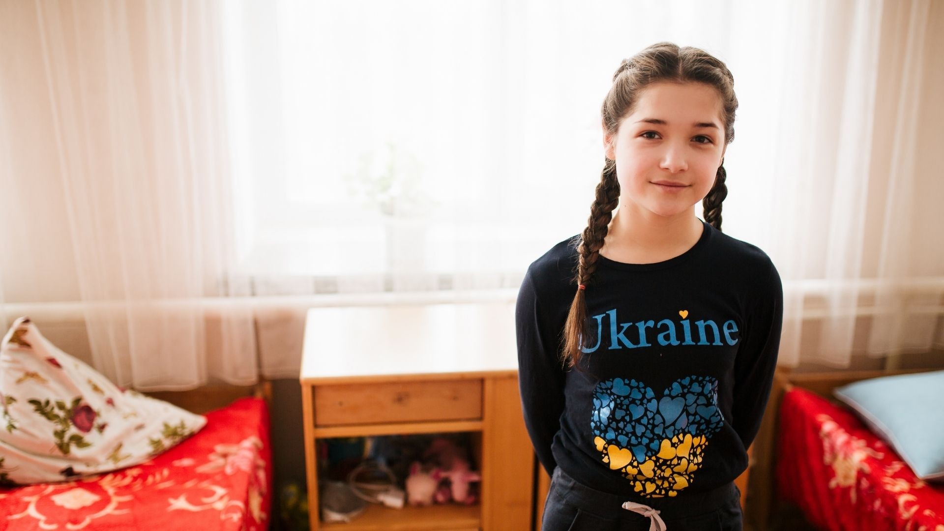 Young girl with braided hair wearing a black Ukraine t-shirt, standing in a dormitory.