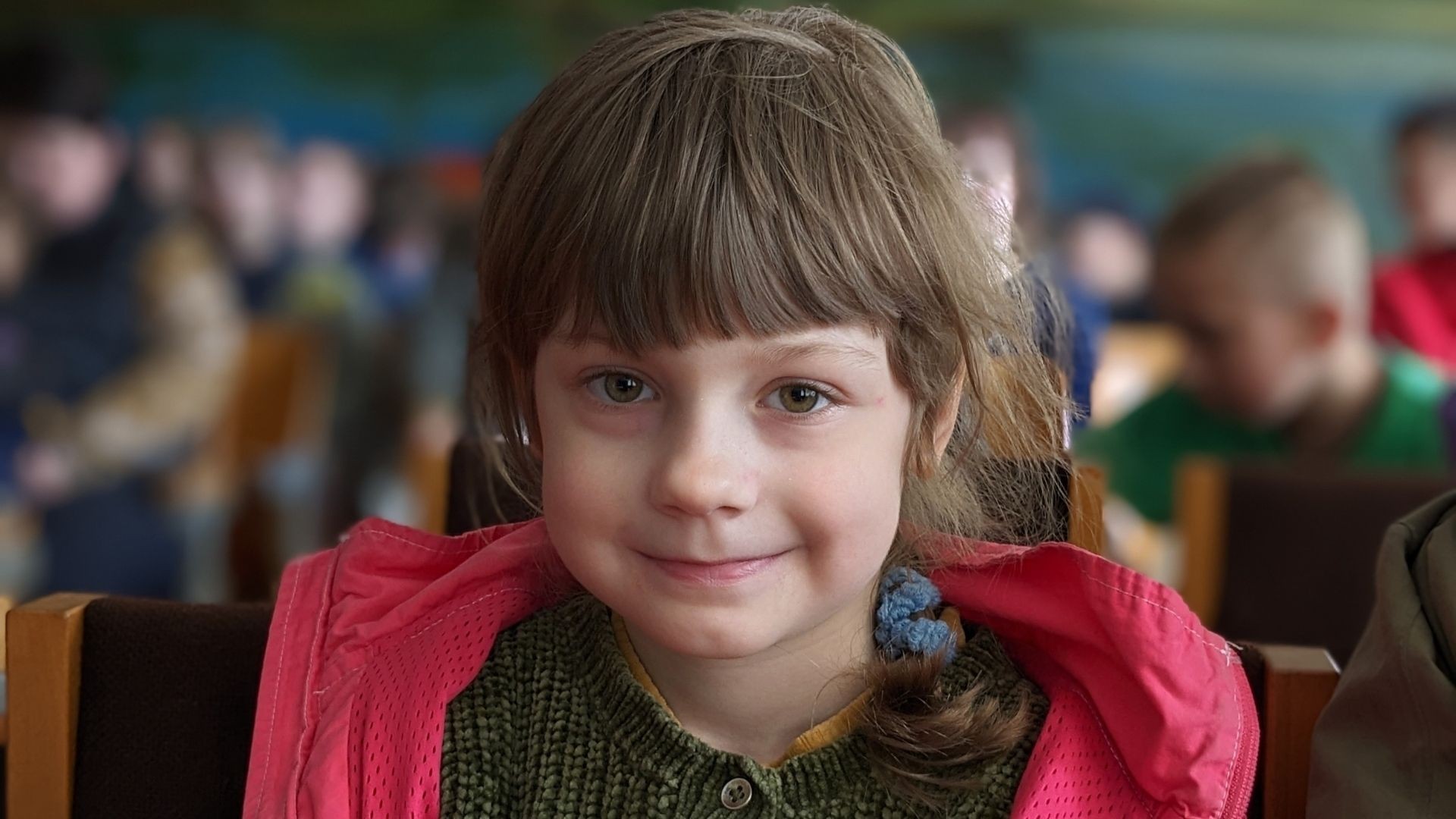Close-up of a young girl smiling shyly while sitting among other children in a crowded room.