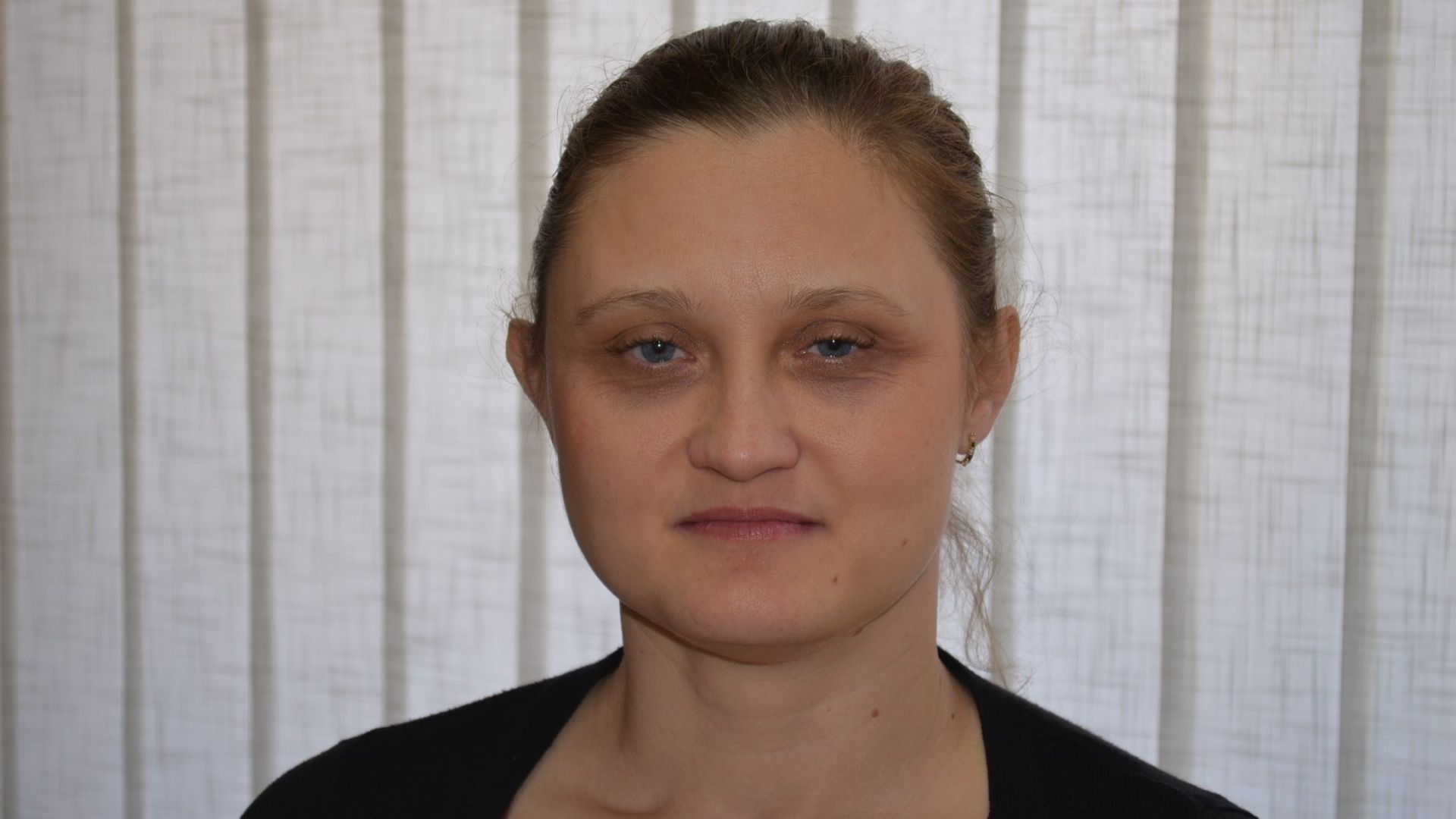 Young woman stands by closed blinds, with a quiet, faint smile.