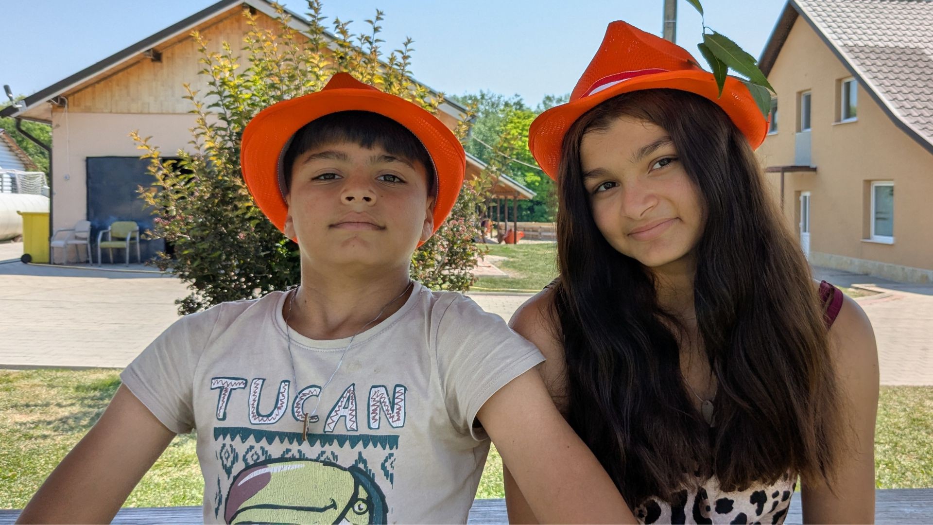 Siblings in orange hats sit together outdoors, enjoying summer camp together
