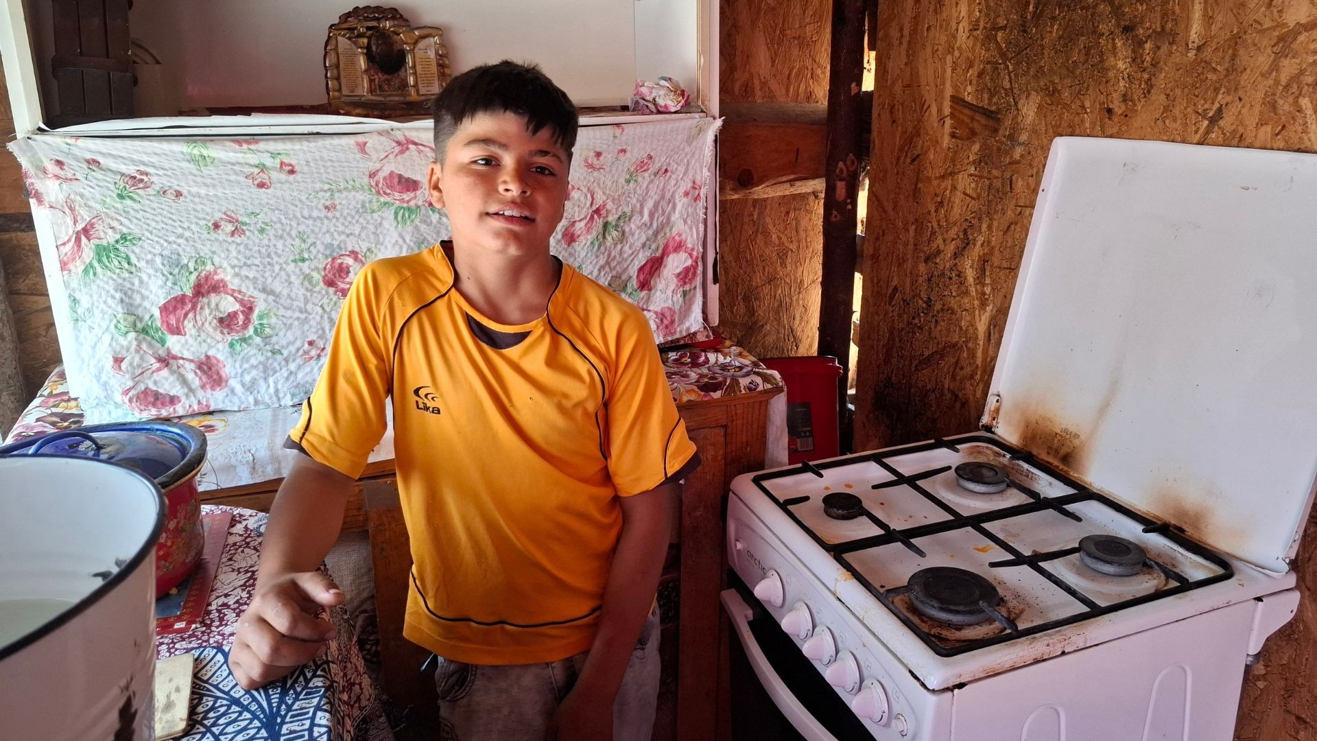 Teen boy in orange shirt stands in cramped kitchen with gas stove and water cans.