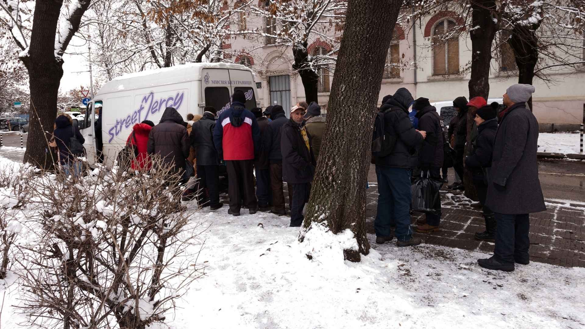A group of people gather around a bus in snowy conditions, waiting to receive food and support services.