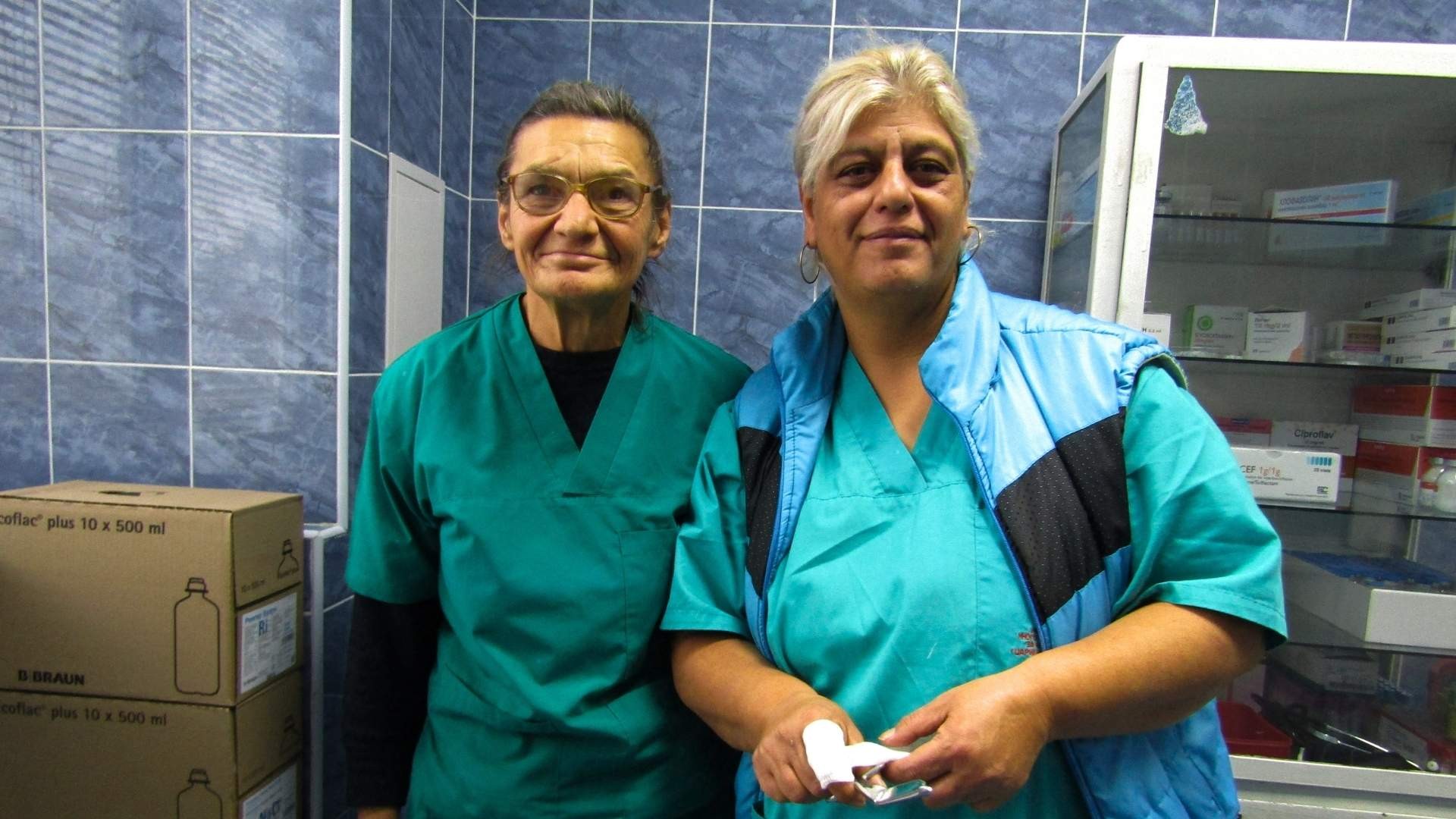 Two women wearing green scrubs stand together in front of a medicine cabinet