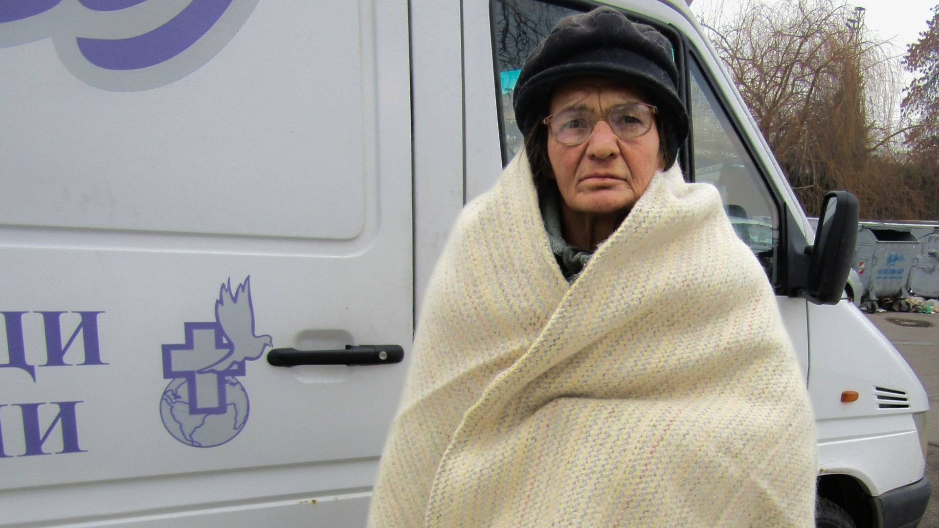 An elderly woman wearing a hat and wrapped in a blanket with a grave expression on her face.