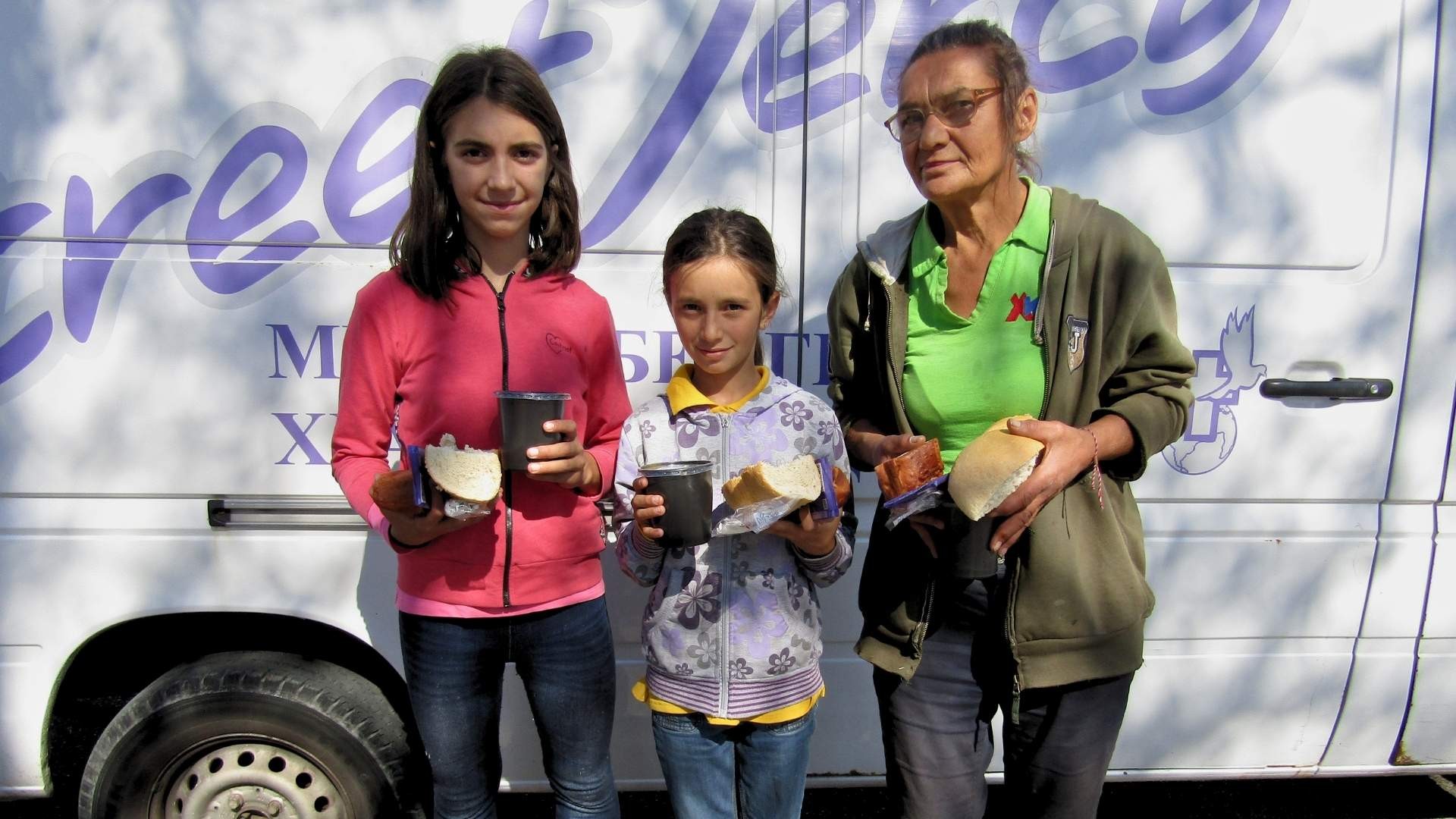 An elderly woman stands with two girls holding food parcels in front of the Street Mercy bus.