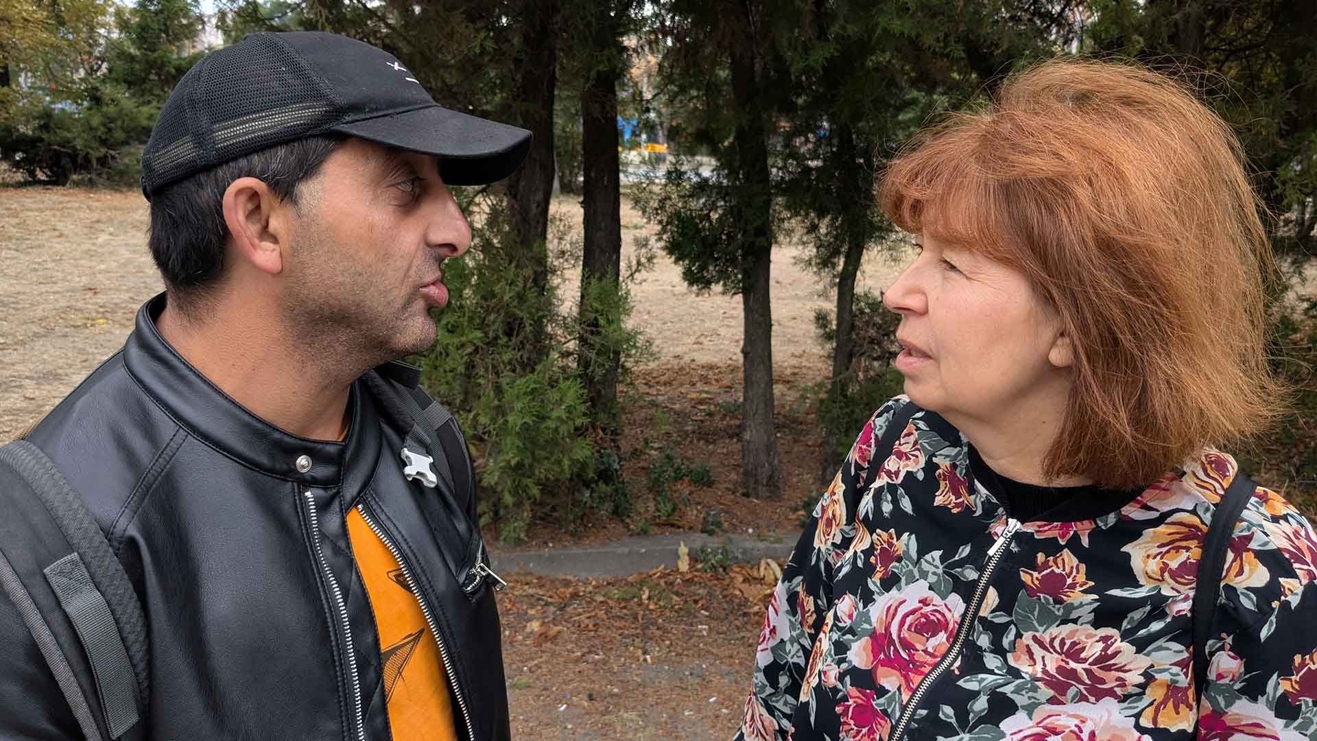 A middle-aged man wearing a cap speaks with a middle-aged woman with red hair during a conversation.