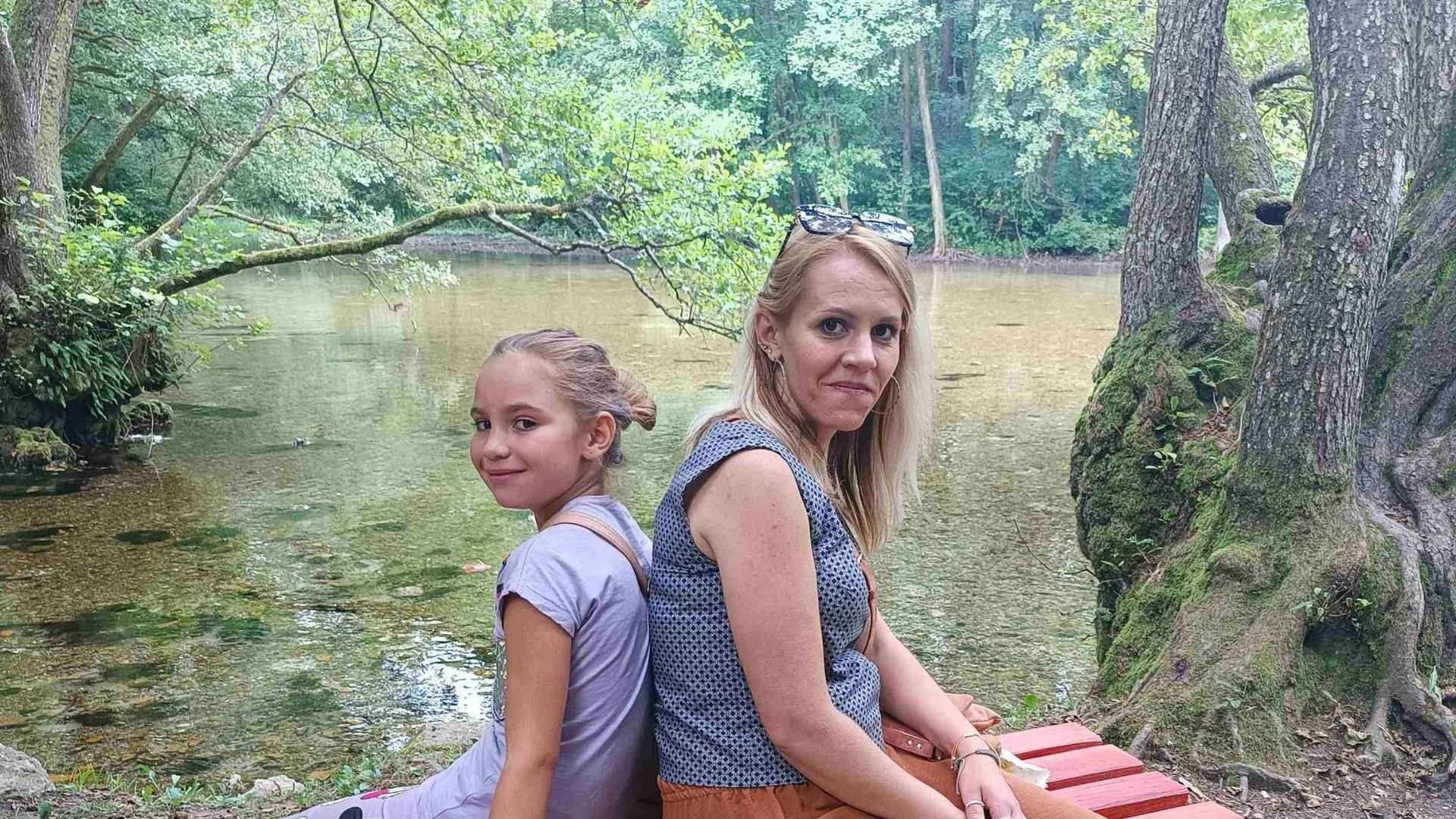 Young girl sits back-to-back with her mother on a bench in lush forest by a spring.