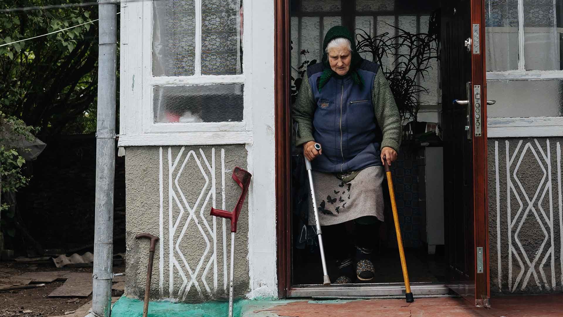 Elderly woman steps carefully out her front door, leaning on a crutch and cane for support.