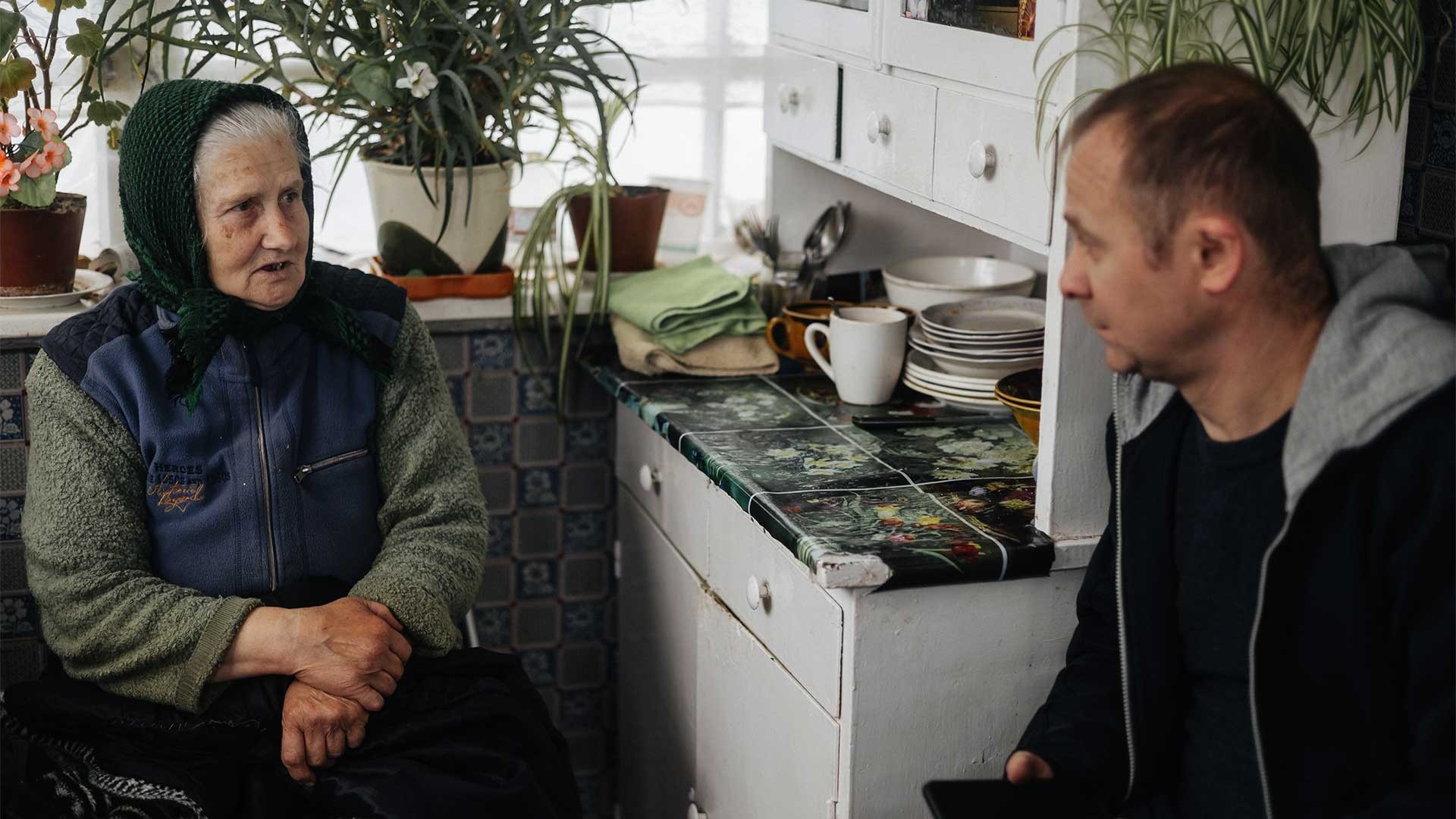 Elderly woman sits in her kitchen, talking with a young man across from her.