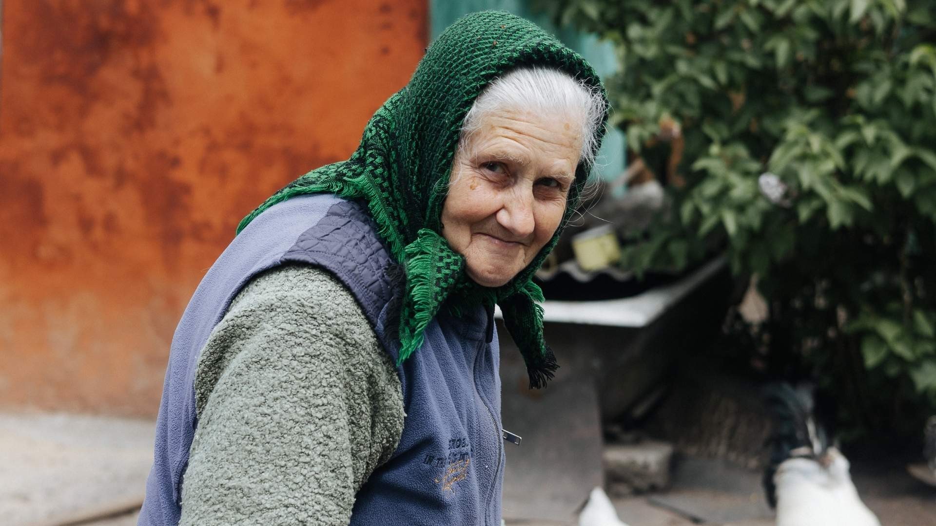 Elderly Nadiya smiles outdoors as she walks to feed her chickens, carrying on daily despite her pain.