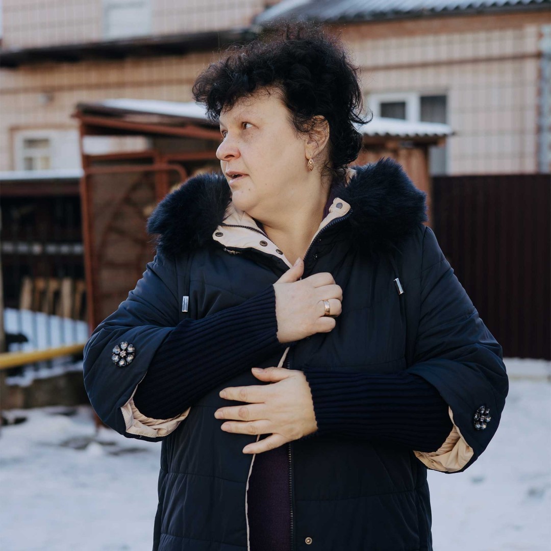 Olena stands in the snow-covered yard, tightening her winter jacket to keep warm.