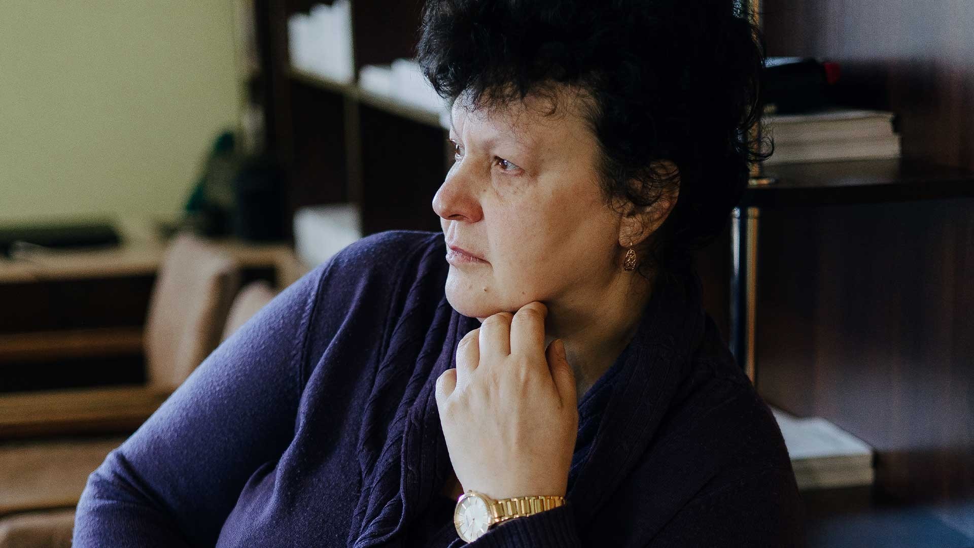Olena sits inside her home, looking sad and weary, lost in deep thought.