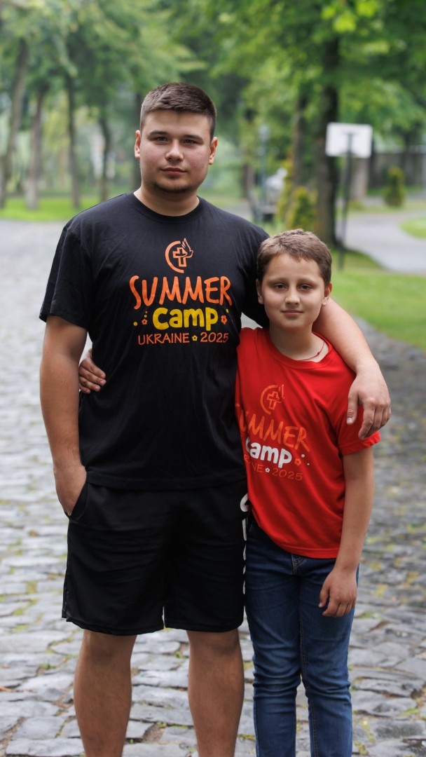 Boy and young camp leader stand arm in arm outdoors at summer camp.