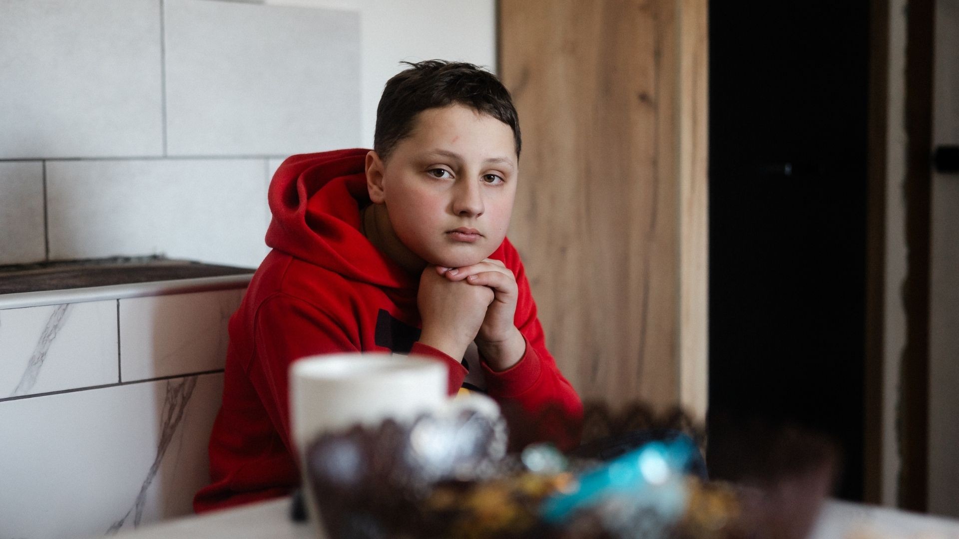 Sad young boy rests his head on folded hands while sitting in the kitchen.
