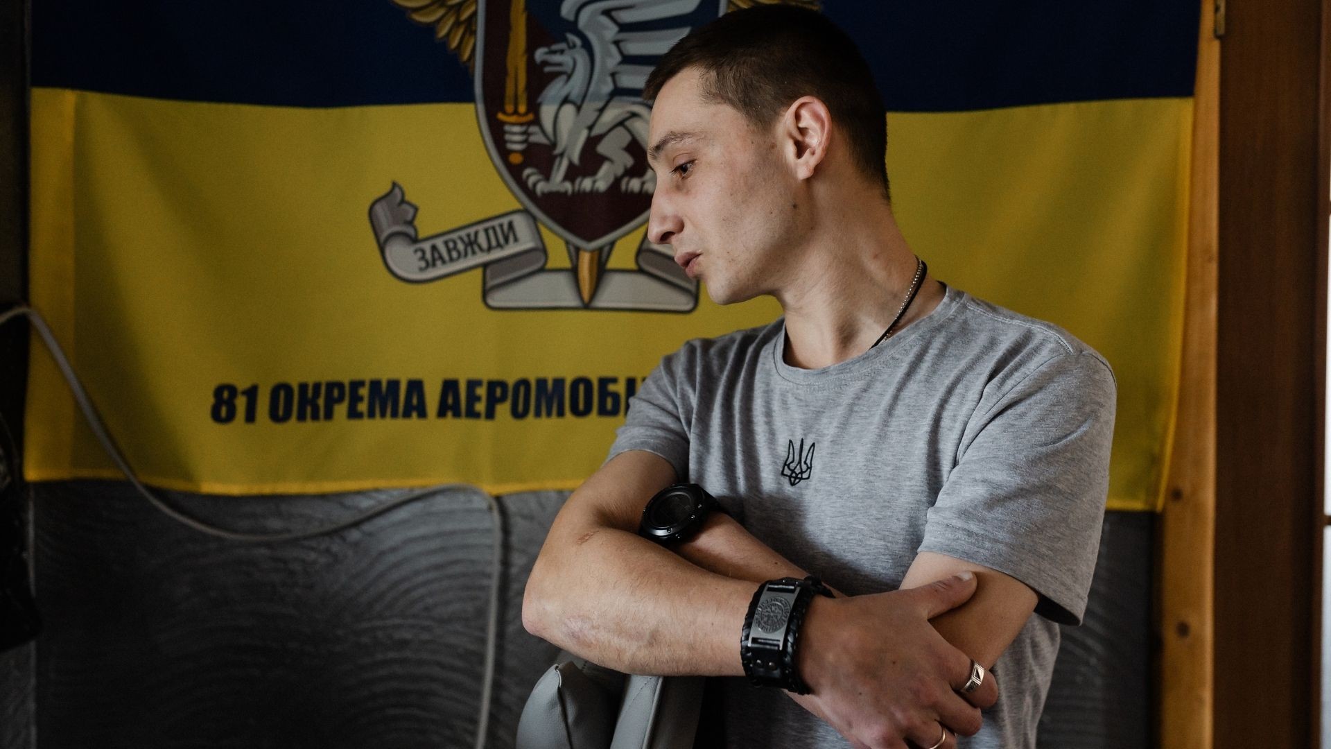 Thin young man stands with folded arms before brigade flag, eyes downcast.