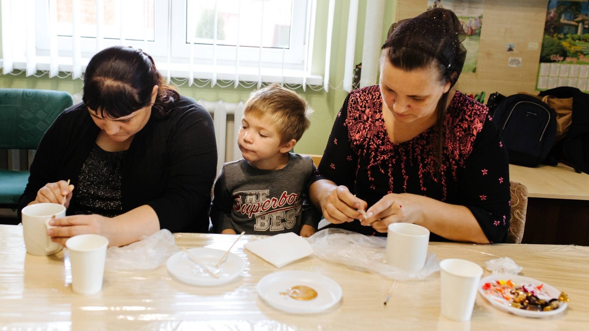 A young boy watches with interest as two women glue and paint broken ceramic cups together during art therapy.