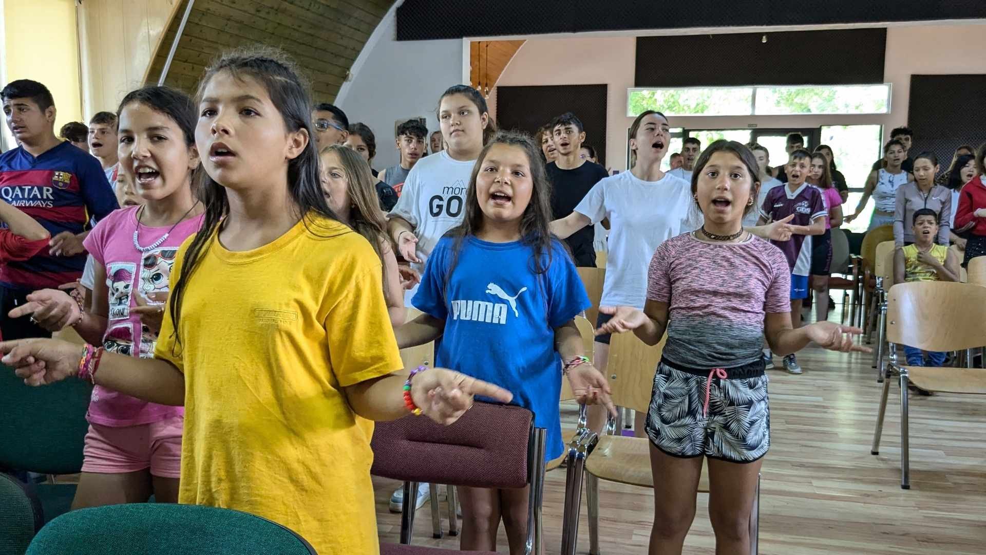 Children standing in a hall, joyfully singing with animated arm movements.